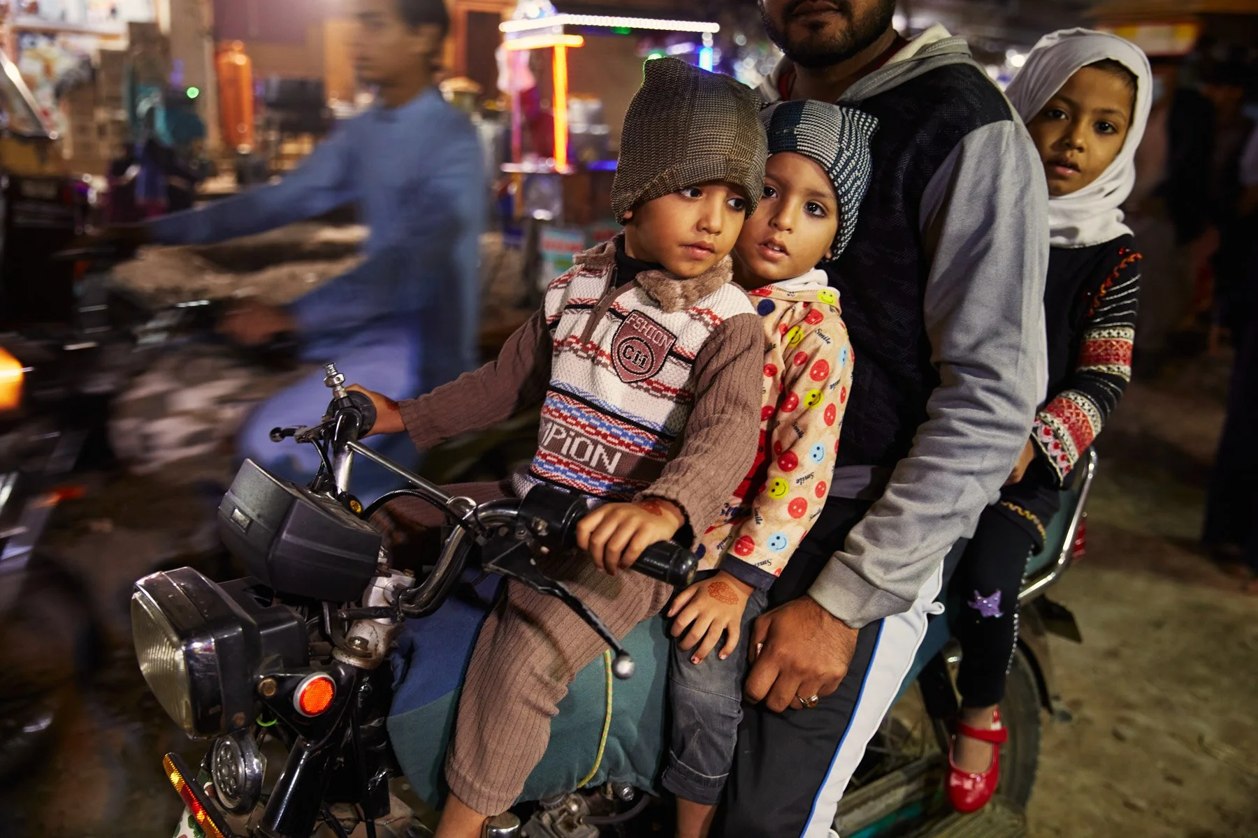 Three children and an adult ride a motorcycle at night in a busy street, with colorful lights and blurred motion of other vehicles in the background. Karachi Pakistan
