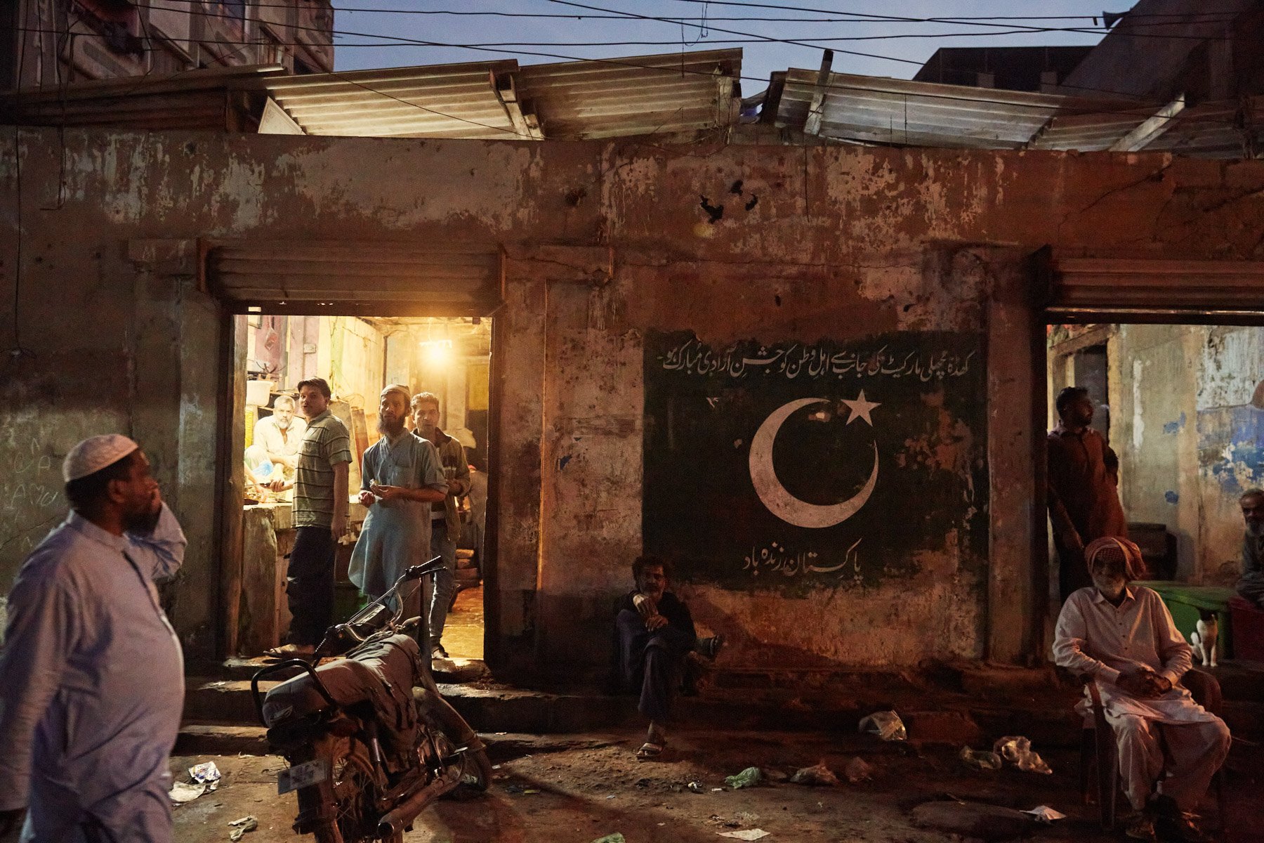 A street scene in Pakistan with men sitting and standing outside a building, some near a motorcycle, and a black flag with a white crescent and star on the wall. The scene appears to be at dusk or night. Karachi Pakistan
