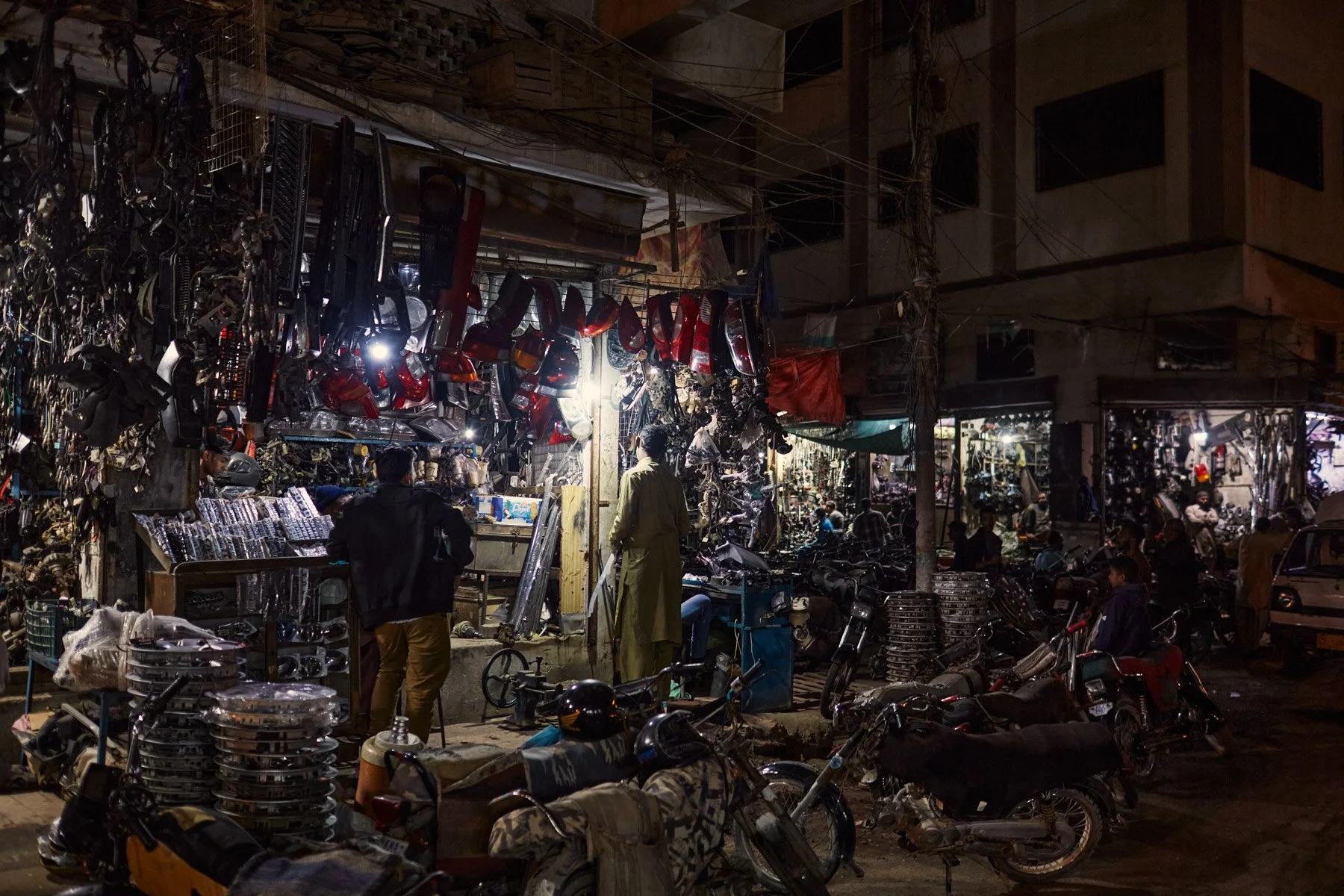 Nighttime scene of a street market with various motorcycle parts and headlights hanging above, people browsing and shopping among motorcycles and car parked on the street. Karachi Pakistan