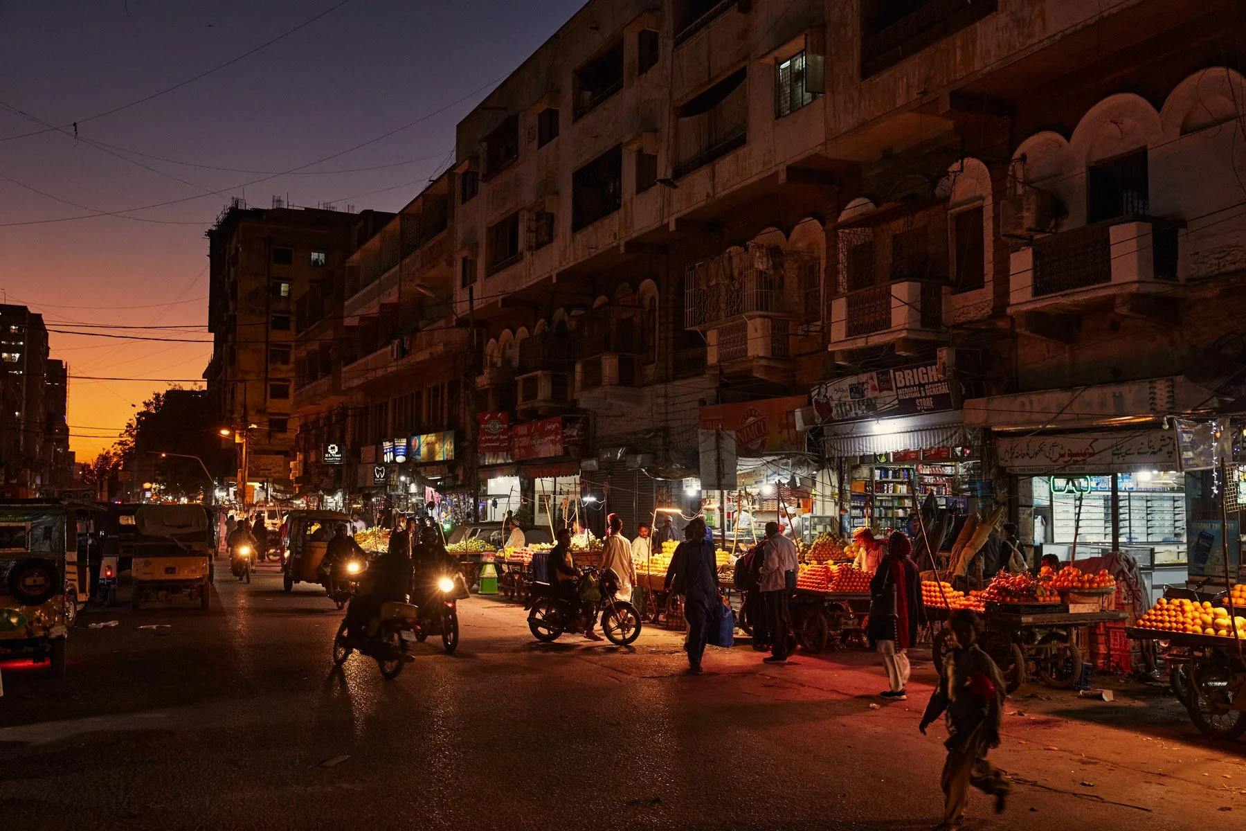 Street scene at dusk with vendors selling oranges and other produce, people walking and riding bikes, motorcycles on the road, and buildings with shops and apartments in the background. Karachi Pakistan