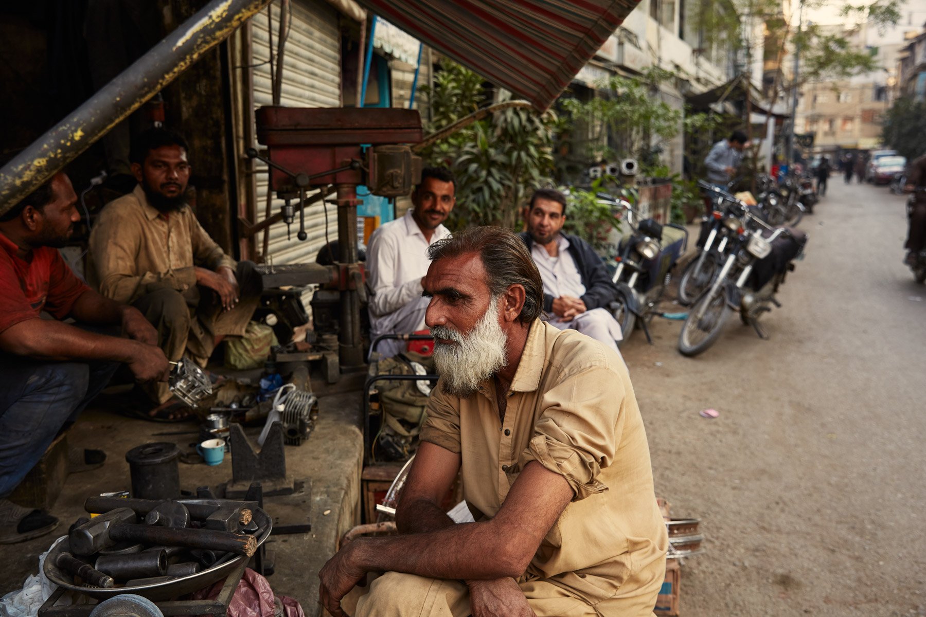 A group of men gathered on a street, with one elderly man sitting in the foreground and others sitting or standing in the background near motorcycles and a small shop. Karachi Pakistan