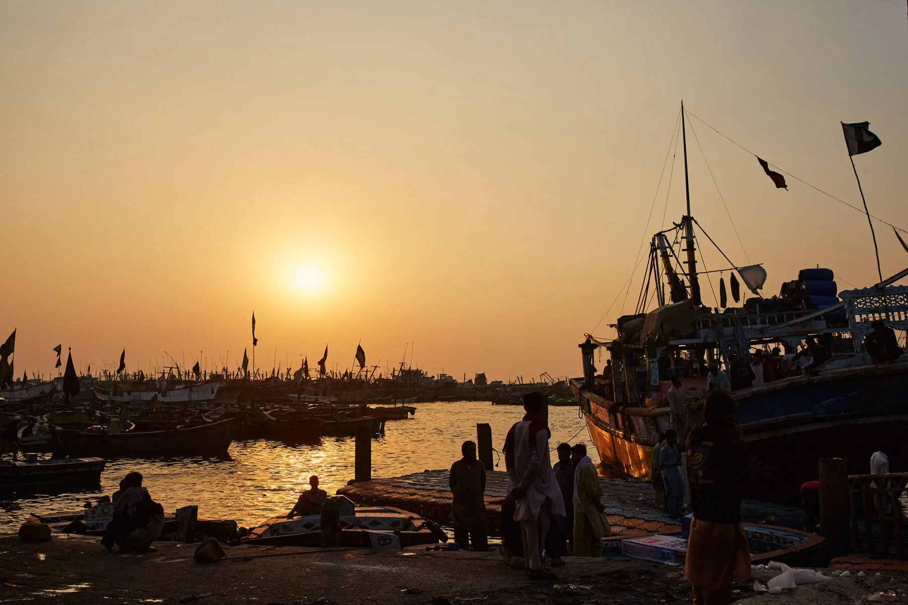 People gather at a harbor during sunset, with boats docked and flags flying at the Dockyard in Karachi Pakistan.