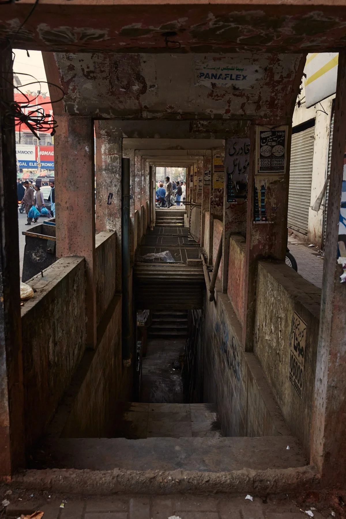 View of a stairwell leading underground, viewed from the top, surrounded by worn, painted concrete walls with posters and advertisements, in a busy urban setting with pedestrians and shops in the background. Karachi Pakistan