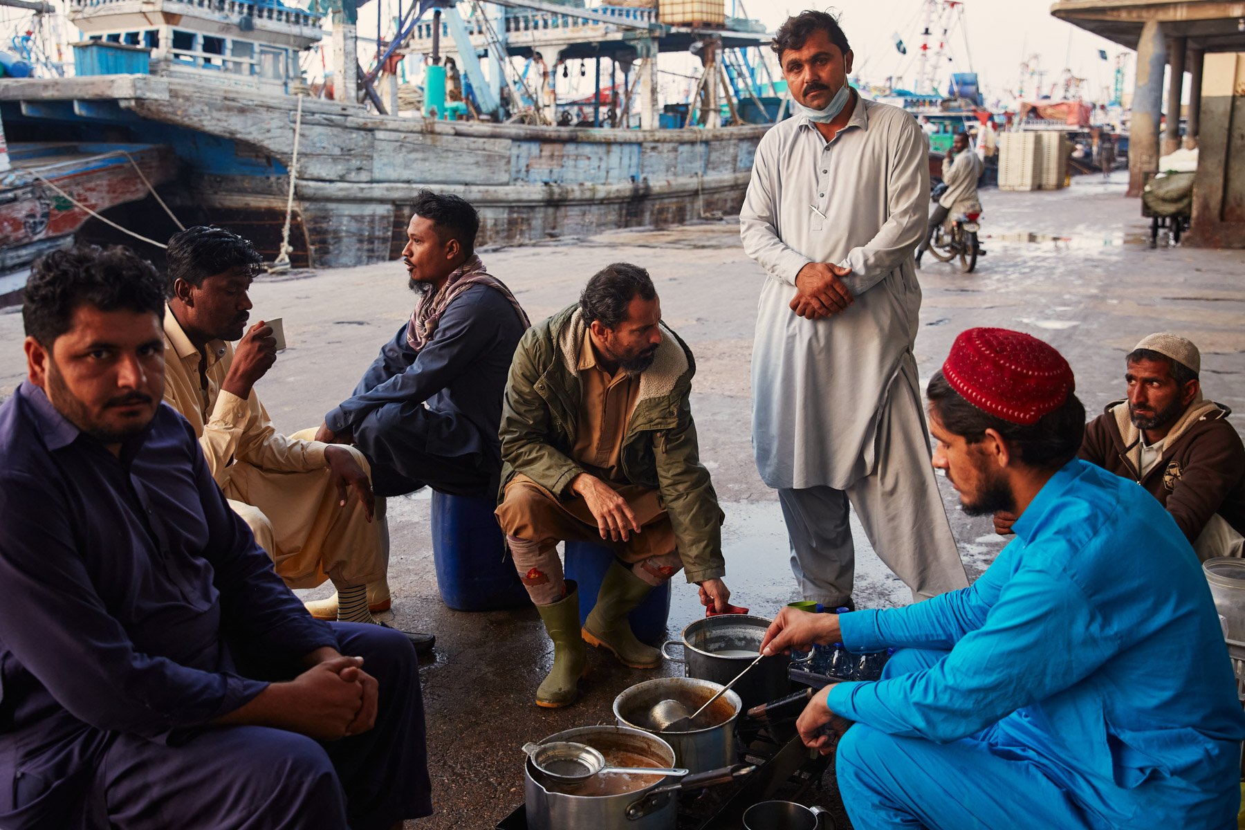 Group of men, sitting and standing, gathered near a street food stall with boats in the background. One man is cooking with pots on a portable stove, others are seated or standing nearby, some wearing traditional clothing. Dockyard Karachi Pakistan