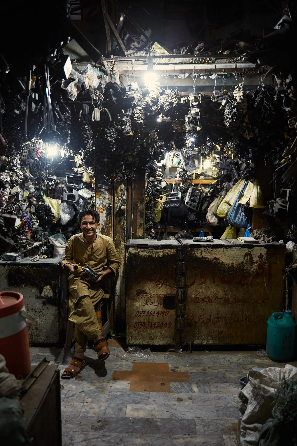A man sitting and smiling in a cluttered shop filled with car parts and electronic components hanging from the ceiling and walls, with bright lights overhead. Karachi Pakistan