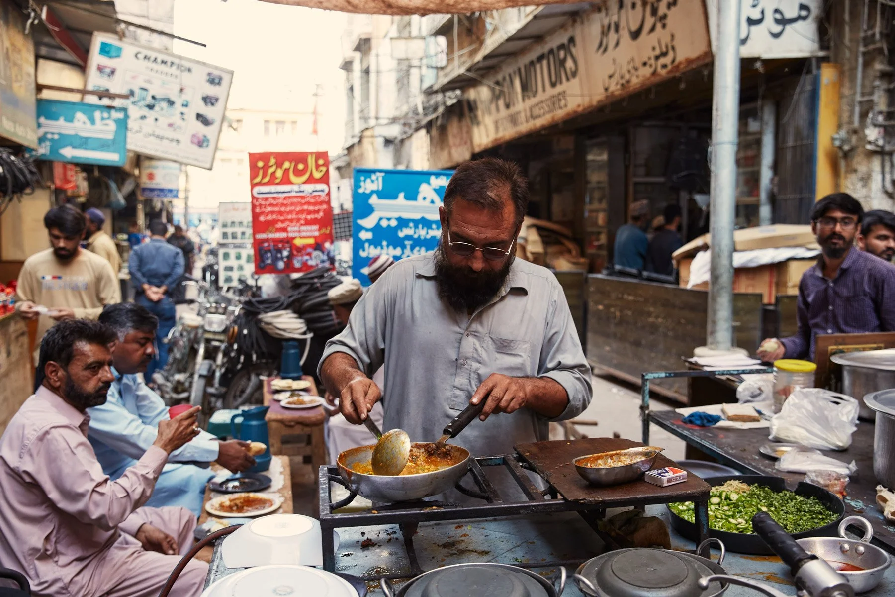 A man wearing glasses and a gray shirt preparing food at a street food stall in a busy market with people sitting and chatting nearby, surrounded by various signs and shop fronts near MA Jinnah Road in Karachi Pakistan.