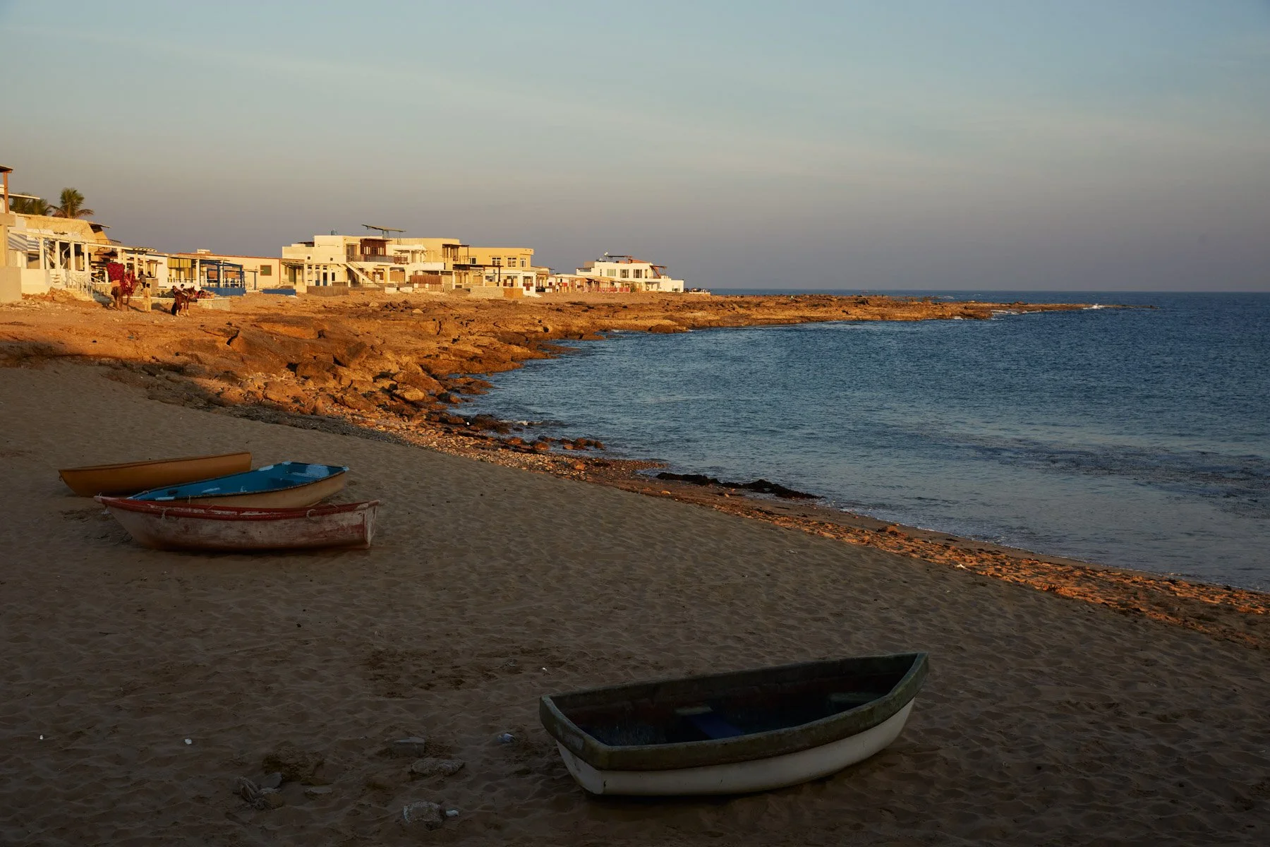 French Beach near Karachi Pakistan, with three small boats on sandy shore, rocky coastline, and modern houses in the distance under a clear sky.