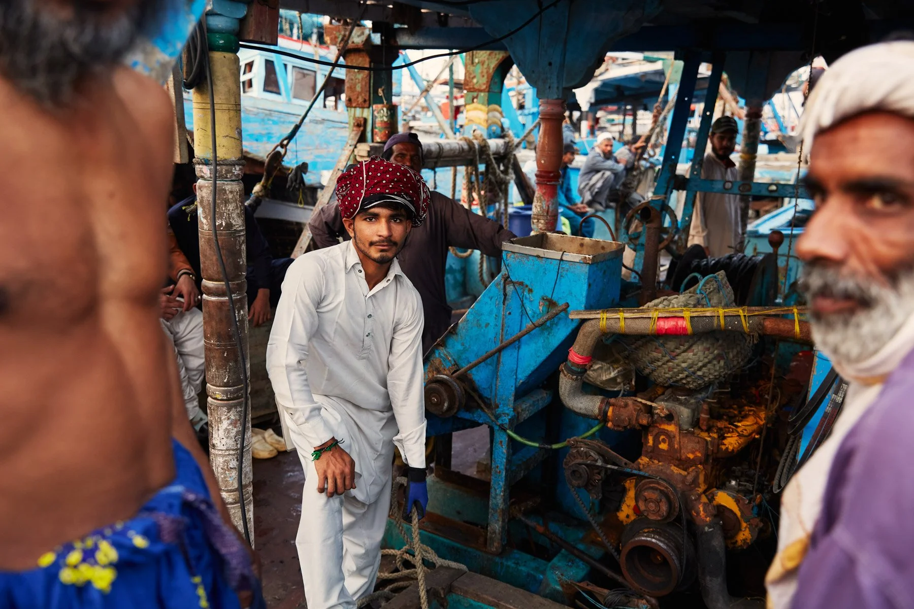 A young man in traditional white clothing and a red head covering standing near a blue boat engine, surrounded by older men and boat parts in Dockyard of Karachi Pakistan.