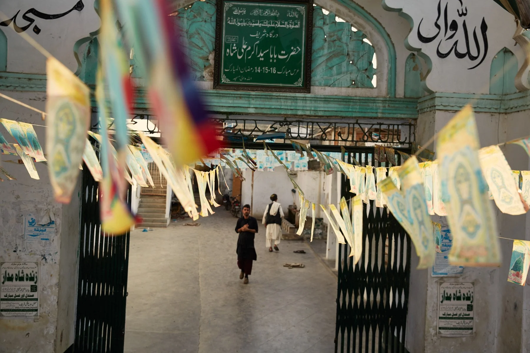 Entrance gate of a mosque decorated with colorful flags, leading into an outdoor courtyard where two men are walking, with a large green signboard in Arabic or Urdu above the entrance. Karachi Pakistan