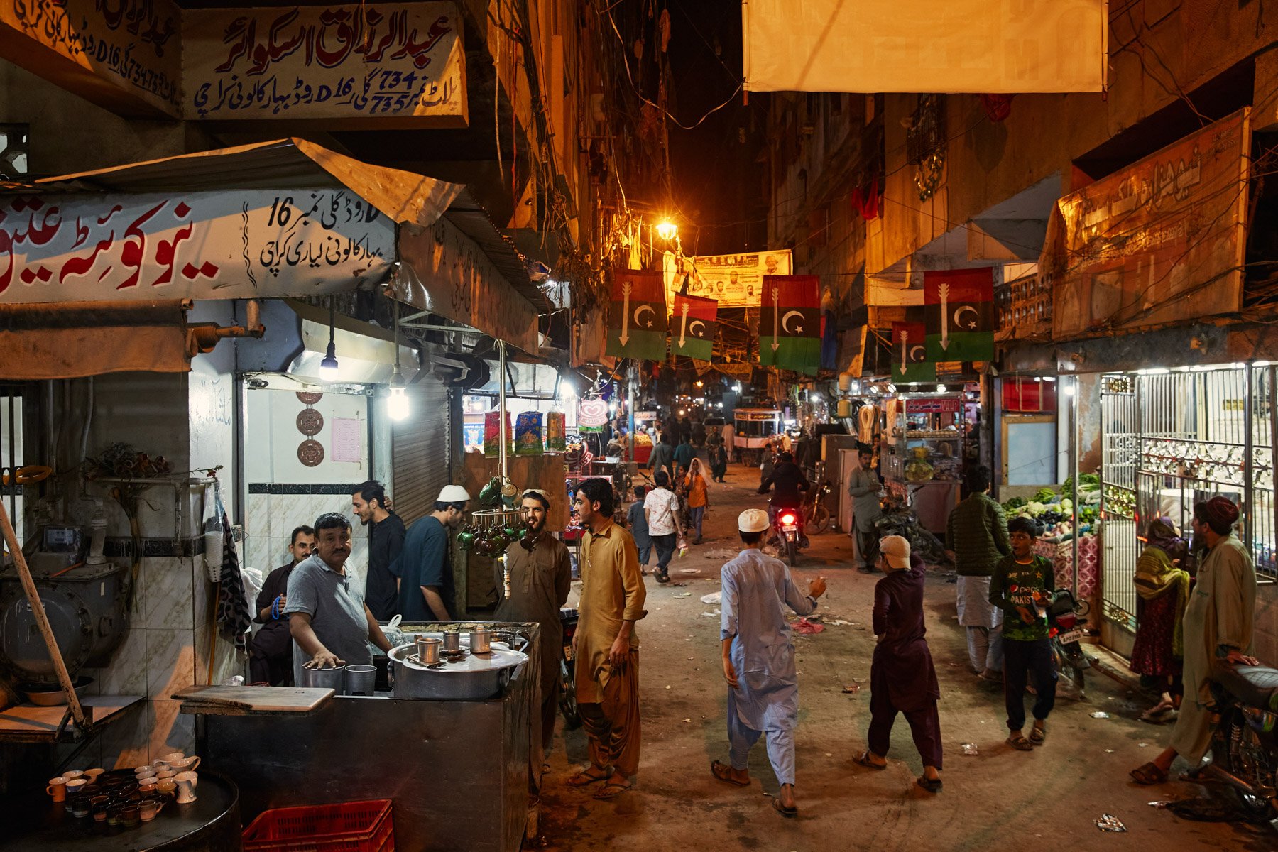 Street market at night with people, small shops, flags, and streetlights, Karachi Pakistan