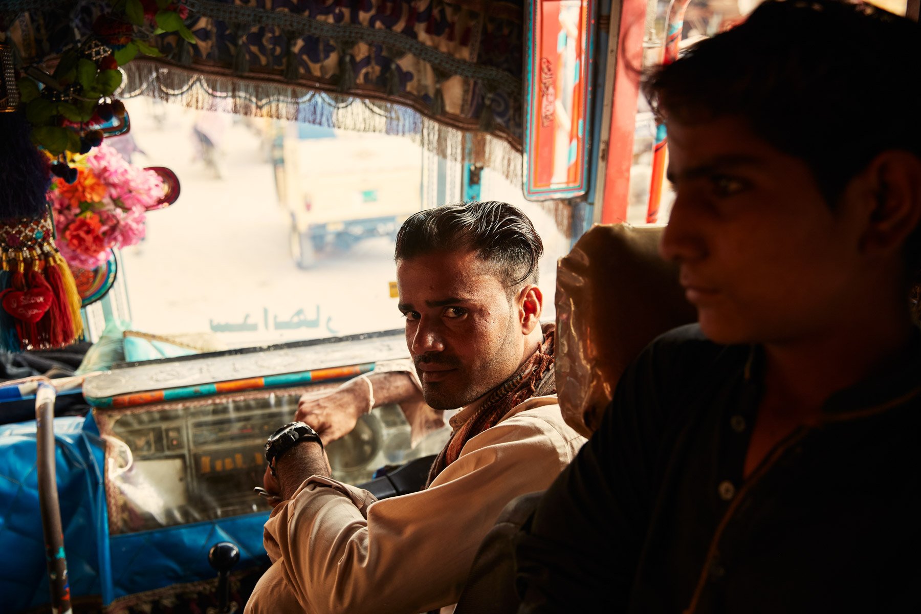 Two men sitting in the front of a colorful bus, with one looking towards the camera and the other in profile, inside a vehicle with a decorative interior in Karachi Pakistan.