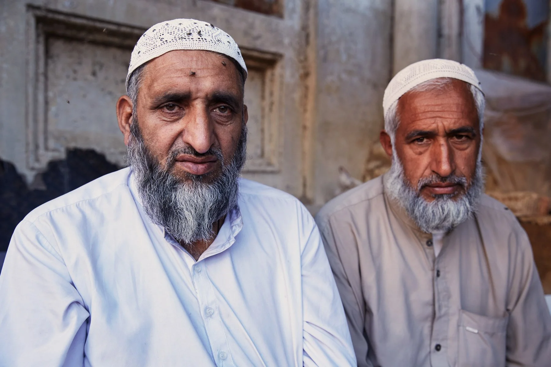 Two elderly men in Empress Market with white beards wearing traditional white caps sit outdoors against a textured wall. Karachi Pakistan