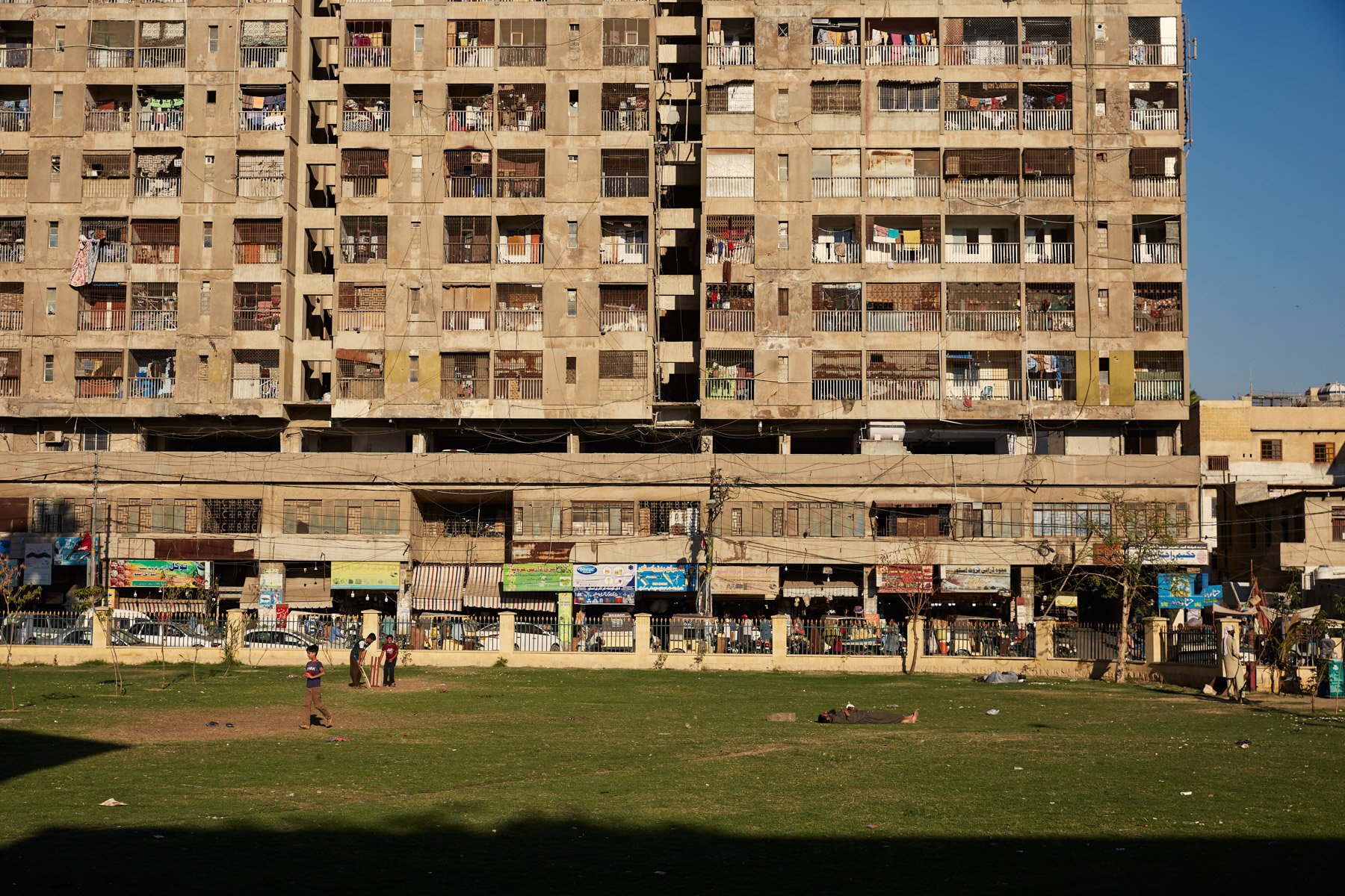Multi-story apartment building with laundry hanging outside, shops at ground level, and children playing on a grassy area in front, near Empress Market in Karachi Pakistan.