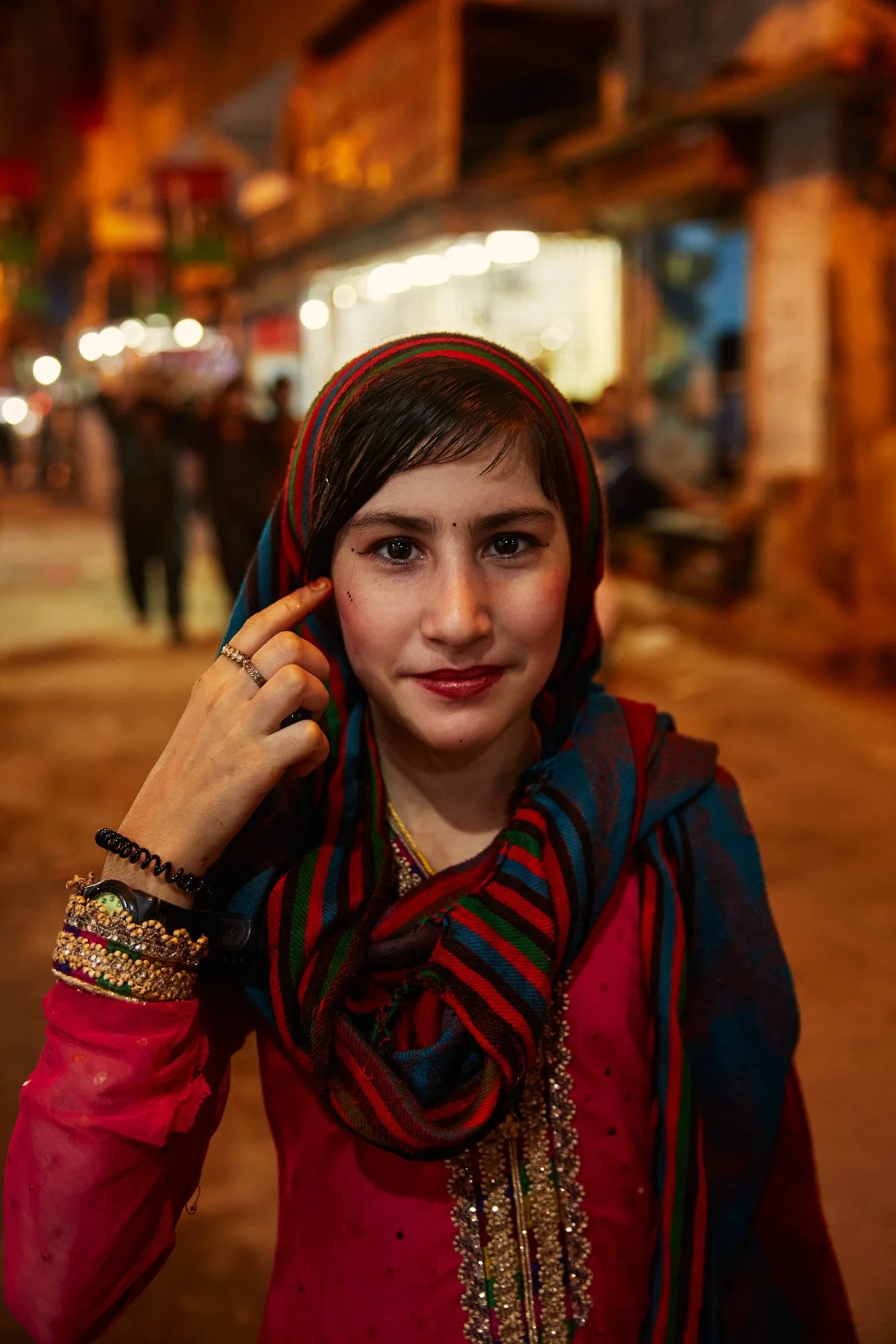 A young girl wearing traditional colorful clothing and jewelry, standing on a lively street at night with blurred lights and people in the background. Karachi Pakistan