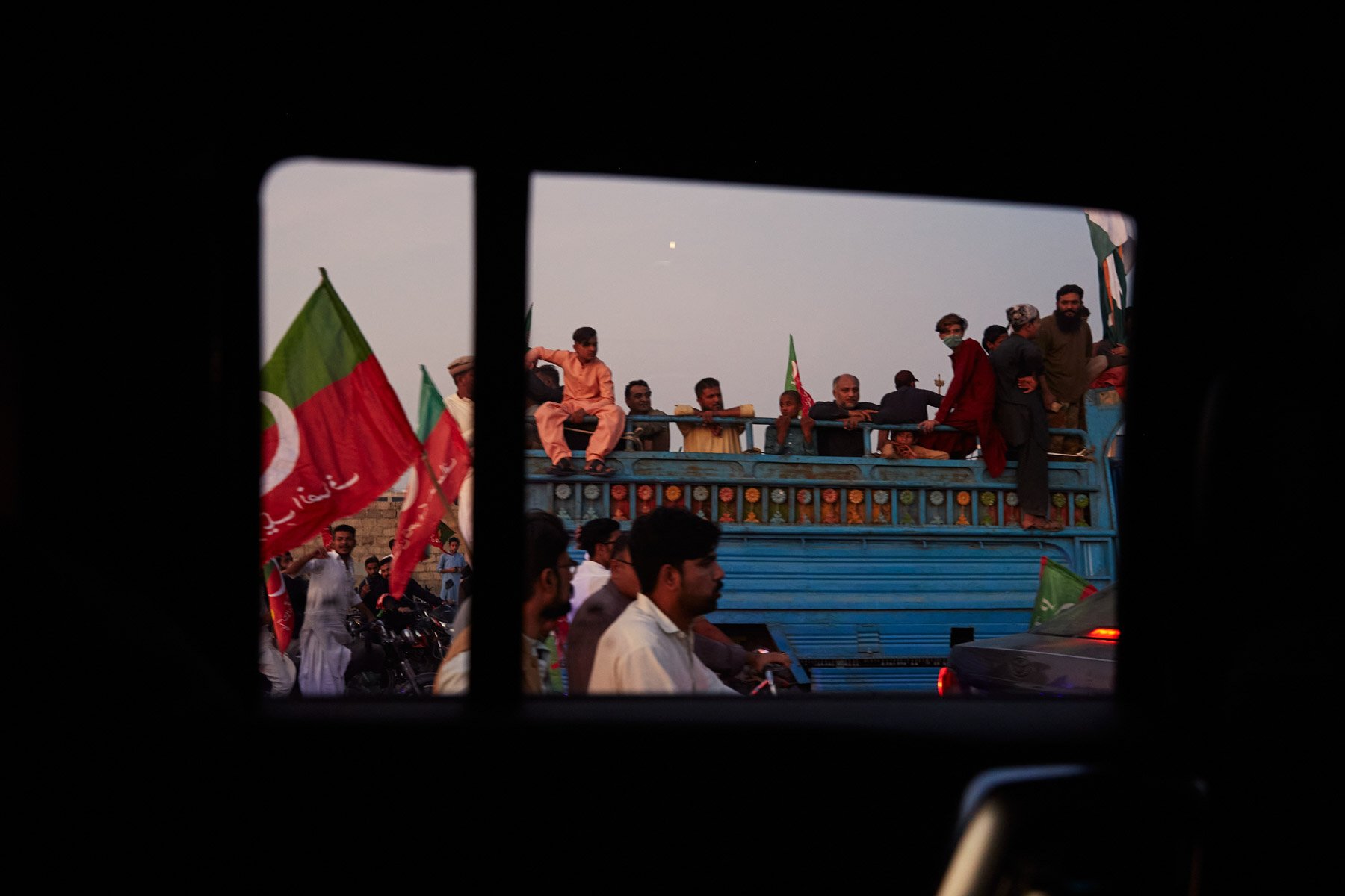 People gathering outdoors, some sitting on a blue truck with a platform, holding flags, with others standing nearby, viewed through a window. Karachi Pakistan