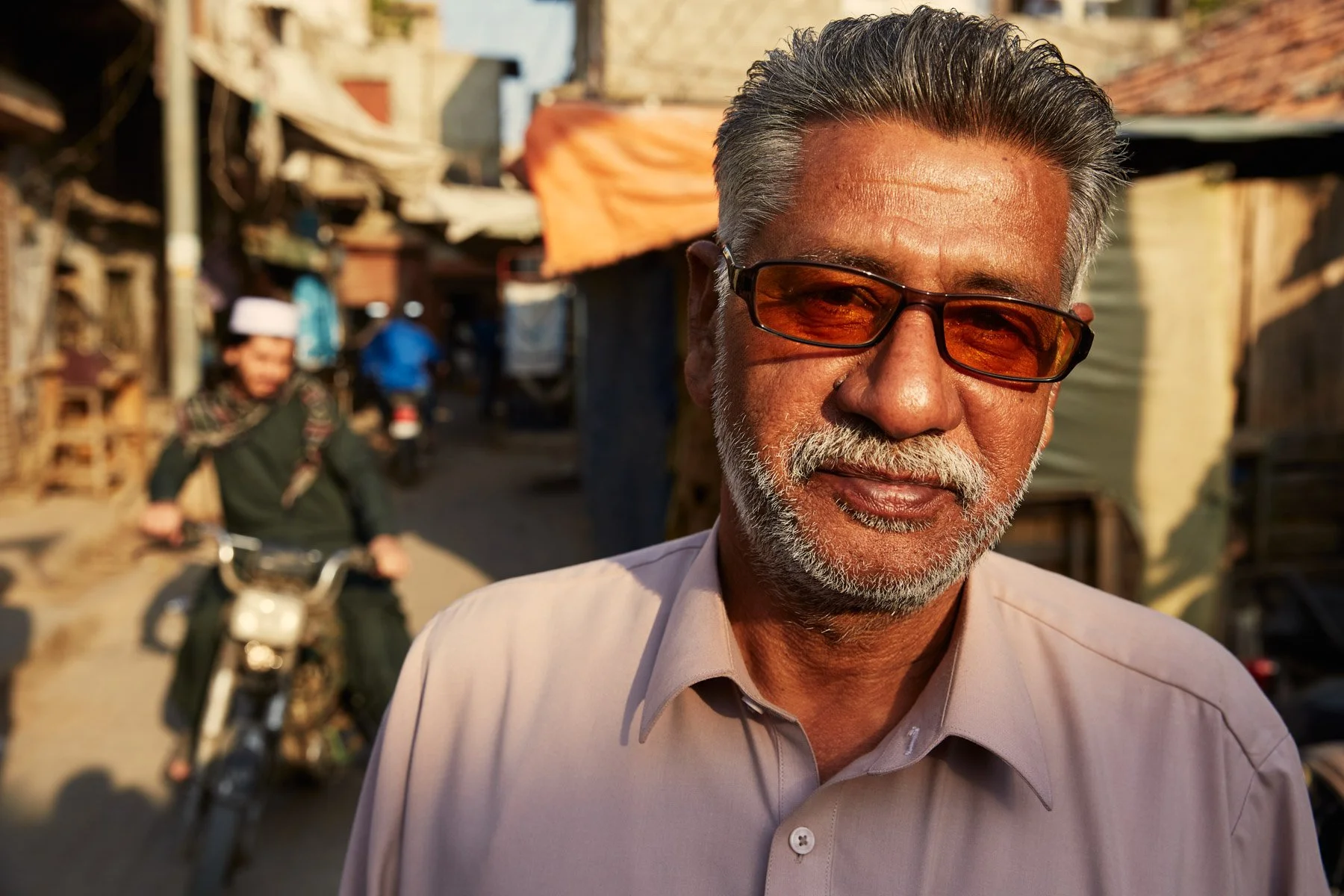 Close-up of a man with glasses and a beard, standing outdoors in a marketplace or street, with a woman riding a bicycle in the background. Karachi Pakistan