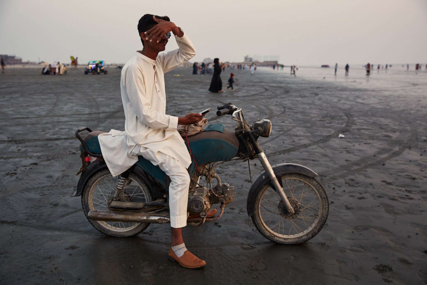 A young man wearing traditional South Asian clothing sitting on a motorcycle on a sandy beach, shielding his eyes and holding a phone, with people and the ocean in the background. Clifton Beach Karachi Pakistan