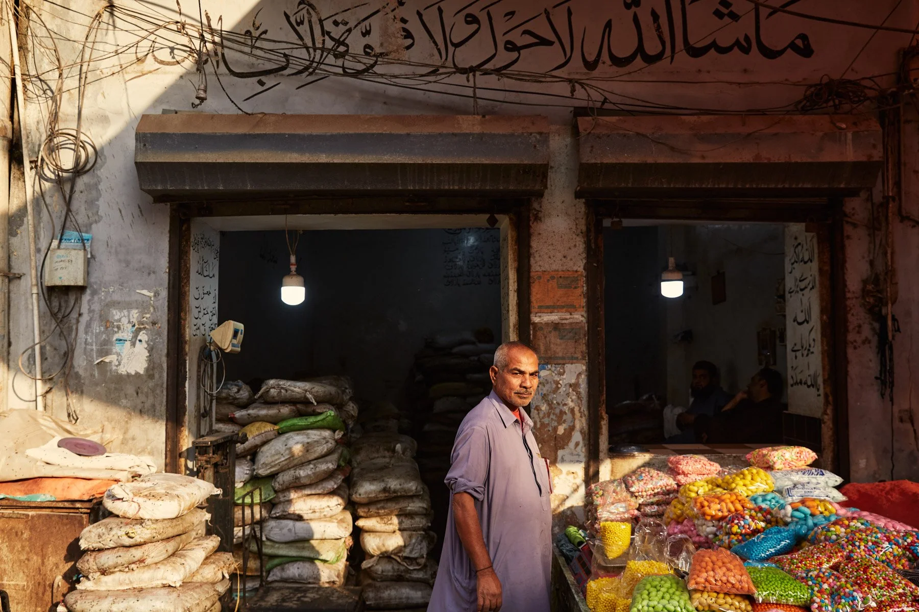 A man stands in front of a street shop selling colorful candies and bags filled with grains or seeds. The shop has stacked bags and is located in a weathered building with Arabic script on the upper wall. Karachi Pakistan