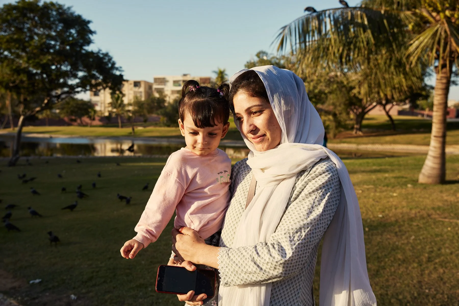 A woman at Agha Khan hospital in Karachi Pakistan, wearing a white headscarf holding a young girl in a pink sweatshirt holding a smartphone in a park with trees and a pond in the background.