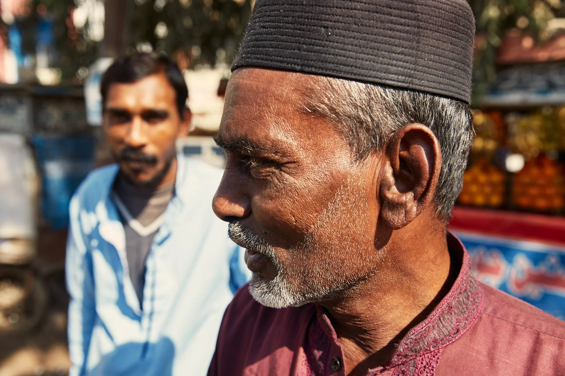 Close-up of an elderly man wearing a black cap, with another man blurred in the background, outdoors in a market or street setting. Karachi Pakistan