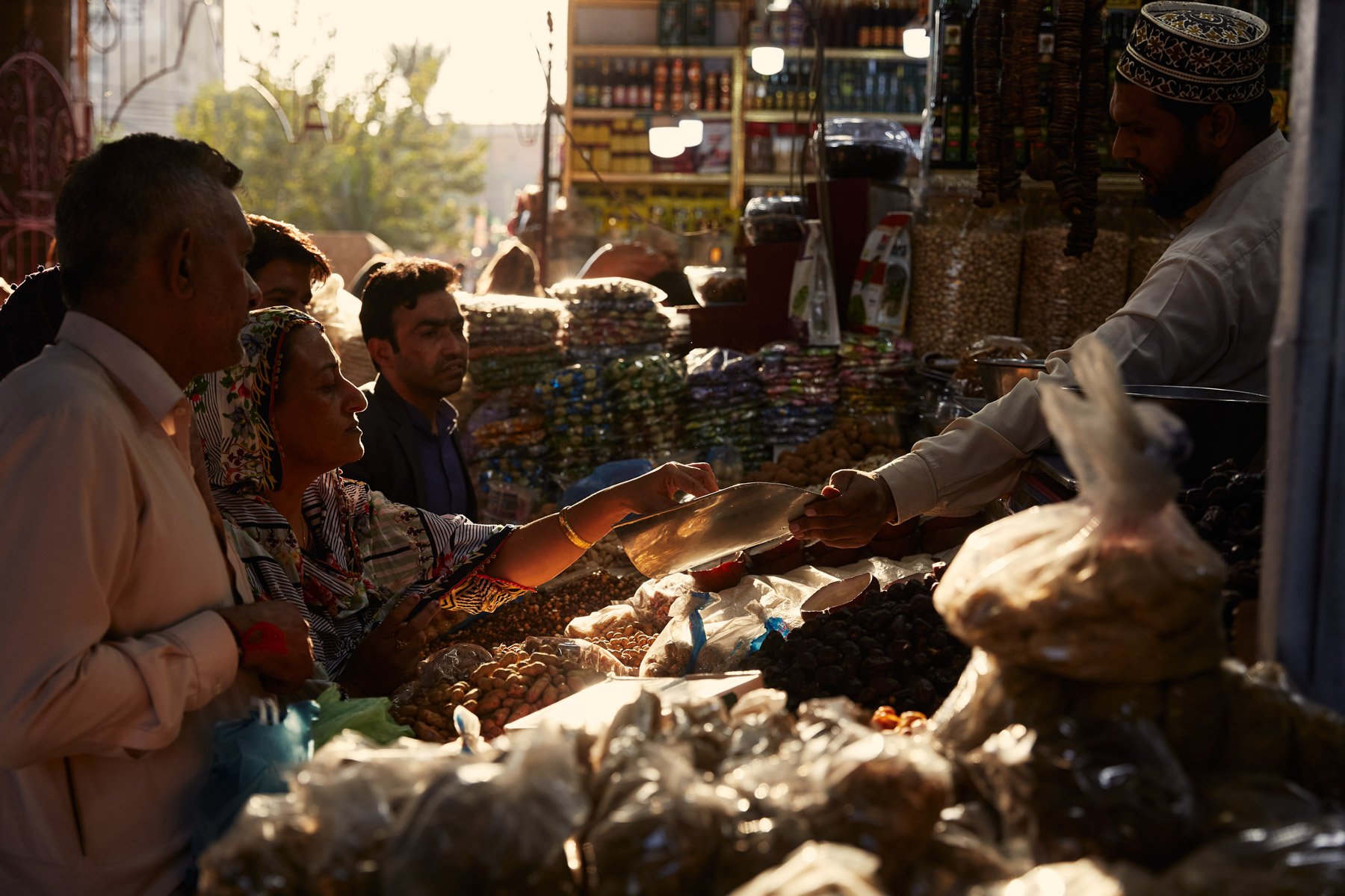 People shopping at Empress Market with various goods, including dried items, packaged and loose products, under warm sunlight. Karachi Pakistan