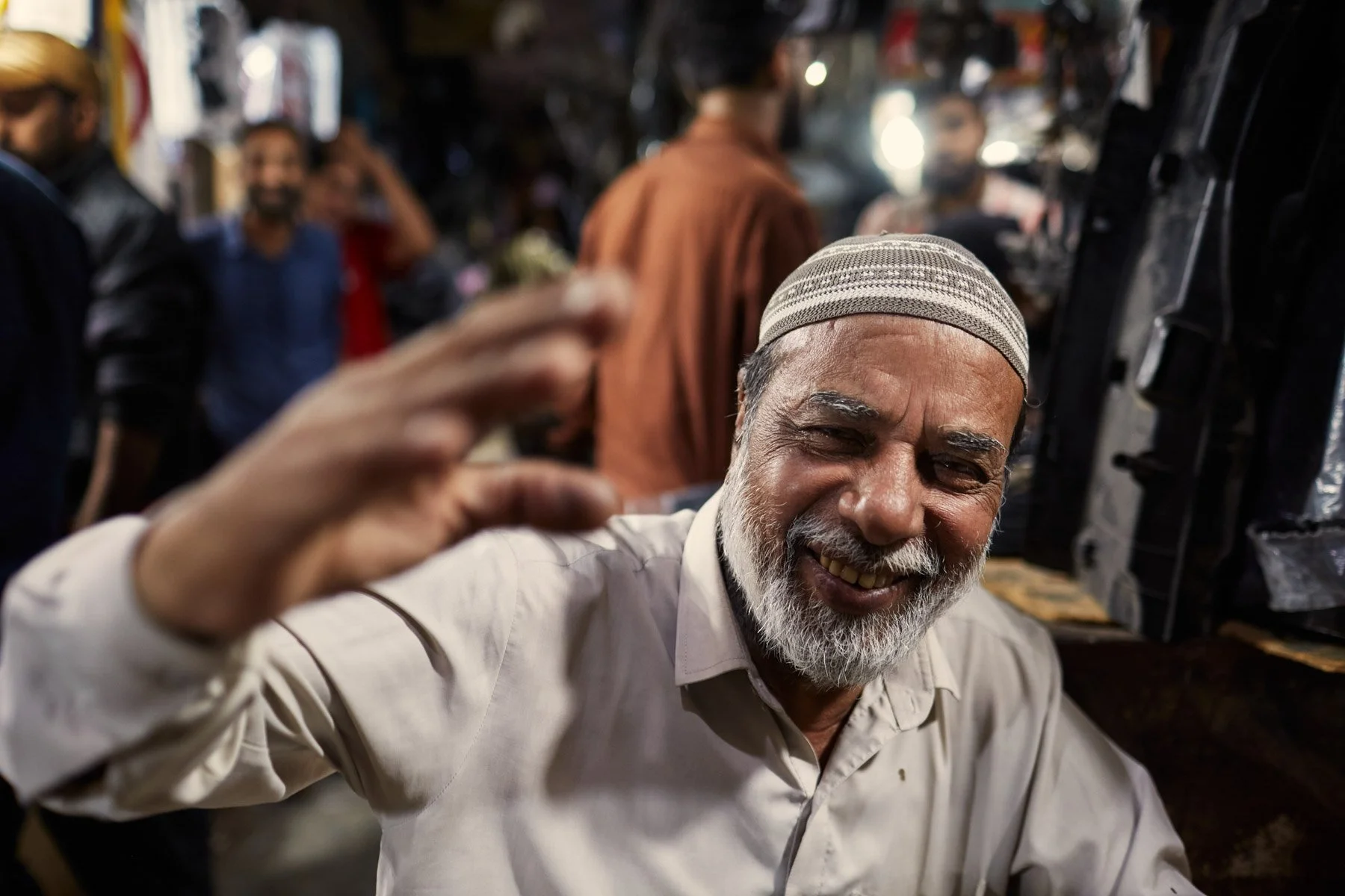 An elderly man with a white beard and a light-colored cap smiling and waving in a busy market or street shop with other people in the background. Karachi Pakistan