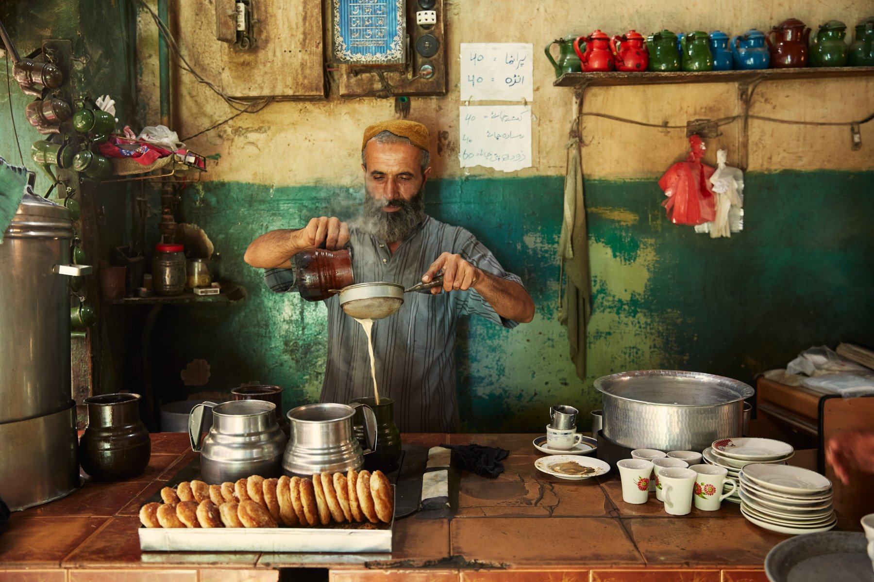 A man with a beard and a brown cap pours tea into a glass from a teapot in a rustic kitchen. The counter has a tray of fried snacks, stainless steel containers, and cups with floral designs. In Karachi Pakistan