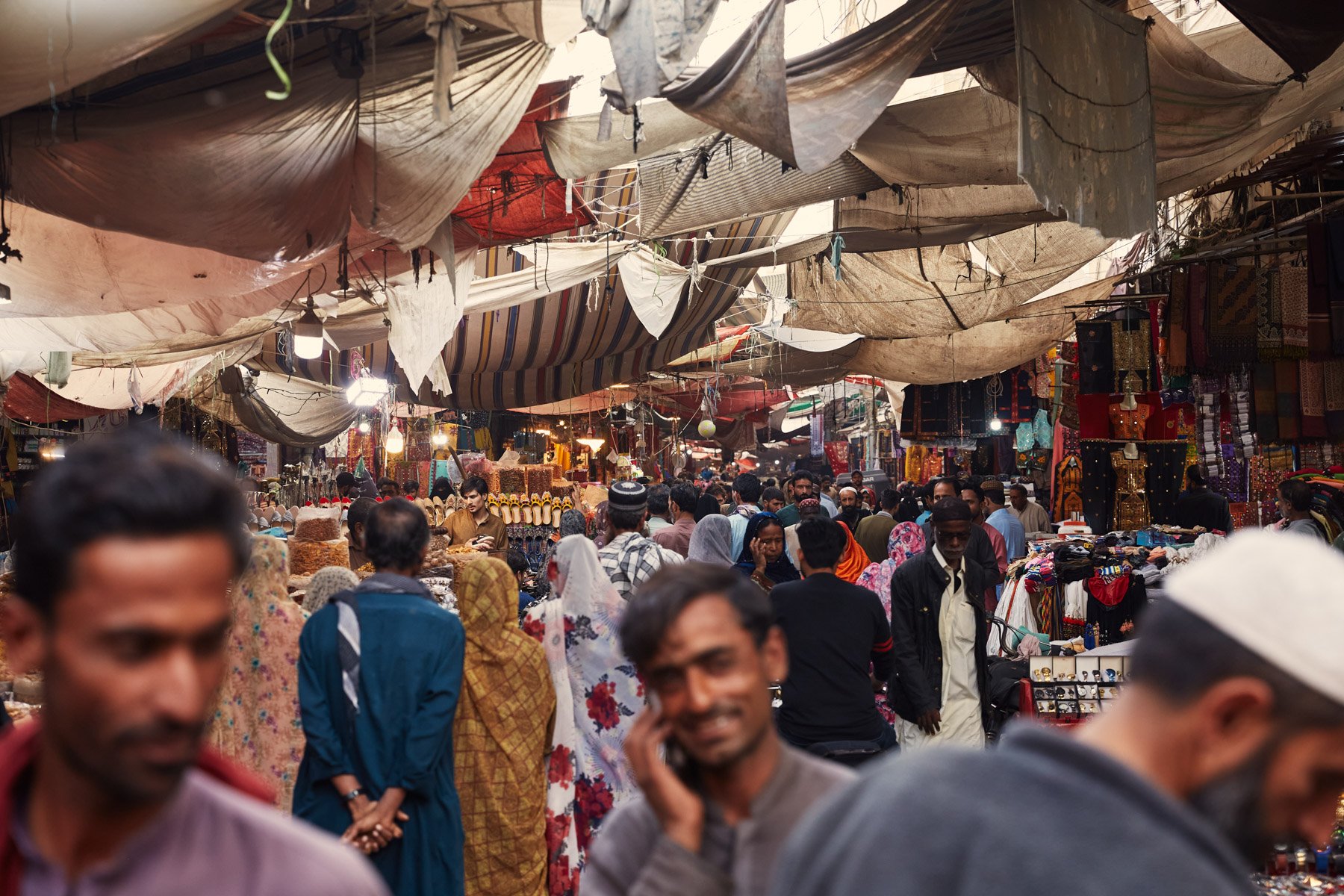 Busy market in an open-air bazaar with stalls selling textiles and clothing, covered with makeshift tarps and fabrics, filled with shoppers and vendors. Karachi Pakistan