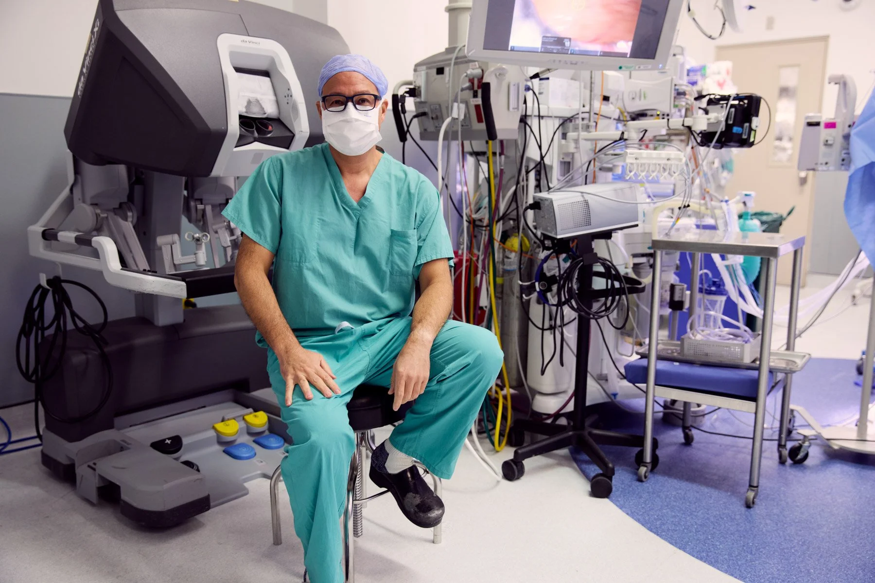 A male surgeon Dr Andrew Kaufman wearing scrubs, glasses, a face mask, and a surgical cap sitting on a chair in an operating room with medical equipment and monitors behind him. At Mount Sinai hospital in New York City