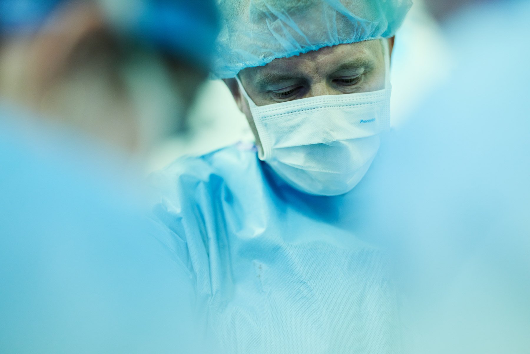 A healthcare professional surgeon wearing a blue surgical gown, mask, and hair cap, focused on a medical procedure. At Mount Sinai hospital in New York City