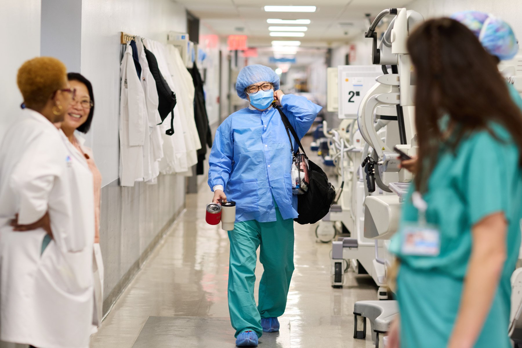 A healthcare worker wearing a blue surgical gown, face mask, and hair cap walking through a hospital corridor, holding drinks and a bag, with other medical staff nearby. At Mount Sinai hospital in New York City