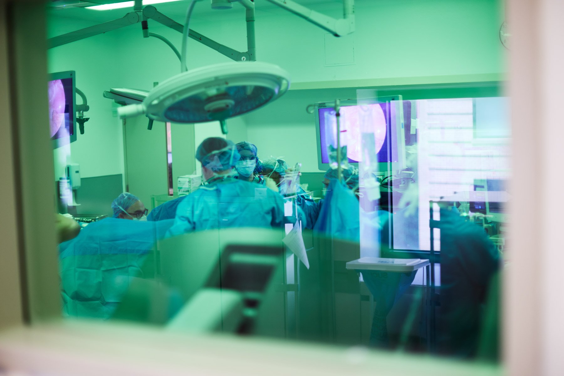 View of an endoscopic gastrointestinal surgical team performing an operation in an operating room, seen through a glass window with reflections, with medical monitors and equipment. At Mount Sinai hospital in New York City