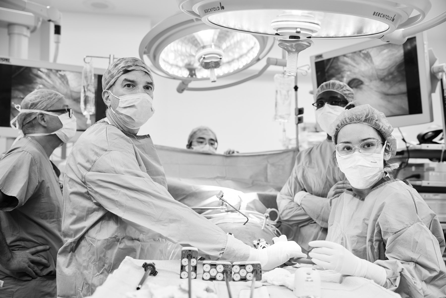A black and white photo of a team of surgeons wearing masks, surgical caps, gowns, and protective eyewear are performing surgery in an operating room. Medical monitors and equipment surround them. At Mount Sinai Hospital in New York City
