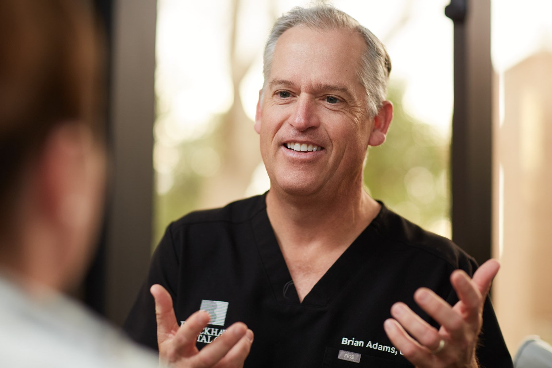 A smiling dentist Brian Adams, wearing black medical scrubs, engaged in conversation with a person whose face is out of focus. The man appears friendly and expressive, gesturing with his hands.