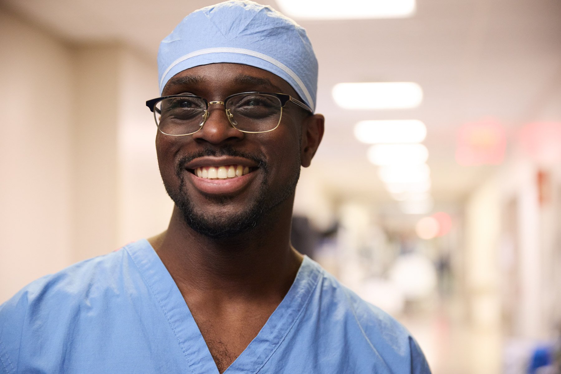 A smiling Black male healthcare professional wearing blue scrubs, glasses, and a surgical cap standing in a hospital corridor. Nigerian surgeon Gbalekan Dawodu. At Mount Sinai Hospital in New York City
