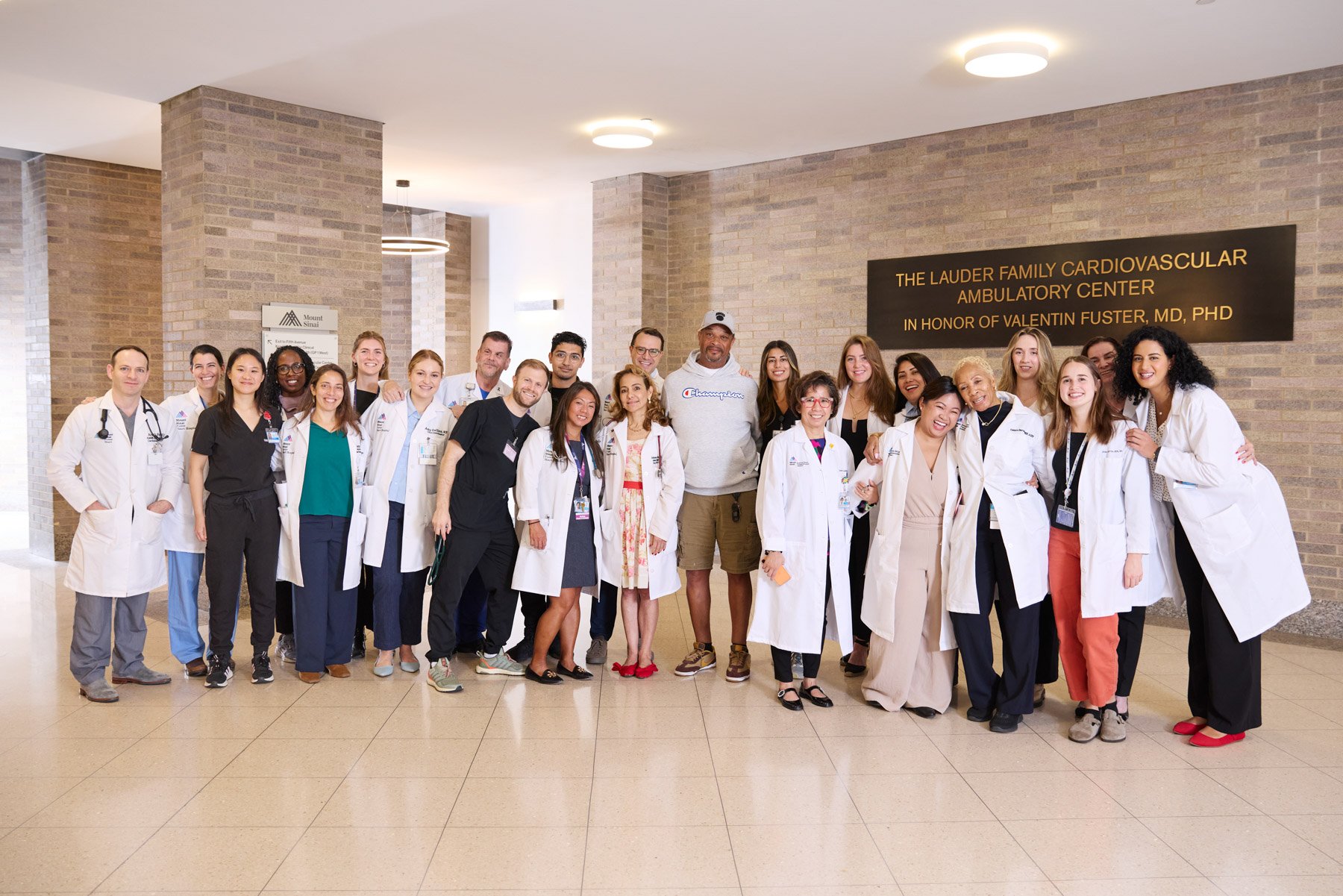 Group of healthcare professionals and staff posing together in a hospital hallway, some wearing white lab coats, in front of a sign that reads 'The Lauder Family Cardiovascular Ambulatory Center in honor of Valentin Fuster, MD, PhD'. At Mount Sinai H