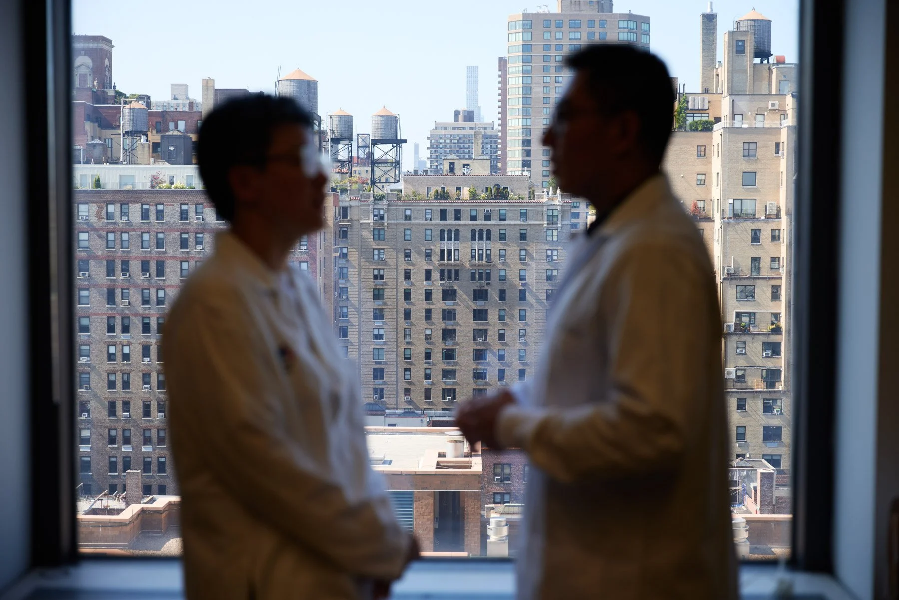 Silhouettes of two scientists Dr Shuang Wang and Dr Xucheng Hou in white coat talking near a window with New York City buildings and water towers outside. At Mount Sinai hospital in New York City