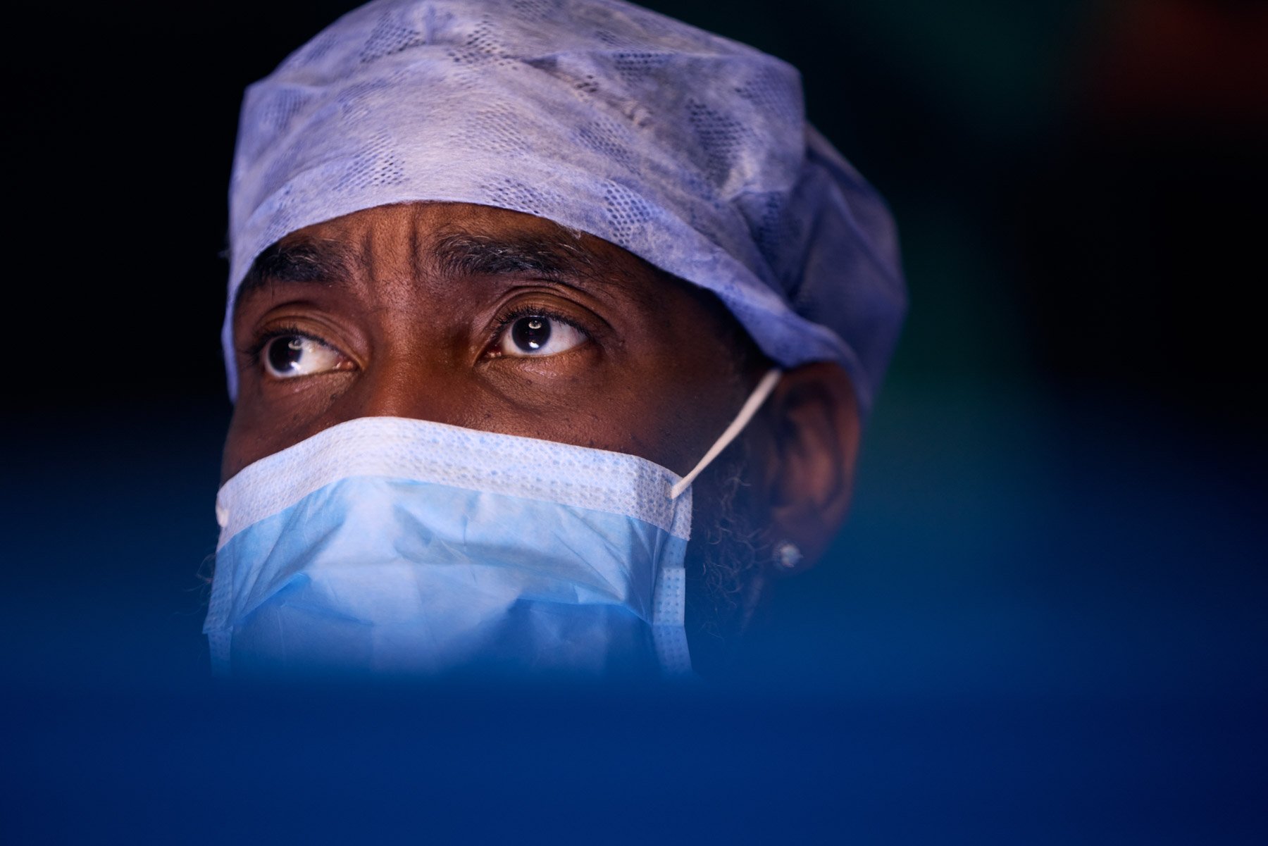 A closeup of a Black man wearing a surgical mask, hair cover, and medical attire, looking upwards. At Mount Sinai Hospital in New York City