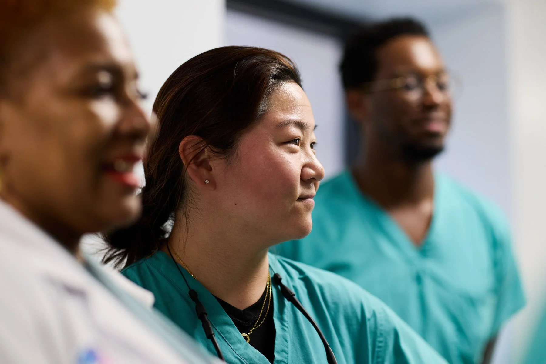 Three healthcare professionals standing side by side, Dr Sue Hahn in the middle, wearing teal scrubs, listening attentively, with one woman in the middle and two men on either side. At Mount Sinai hospital in New York City