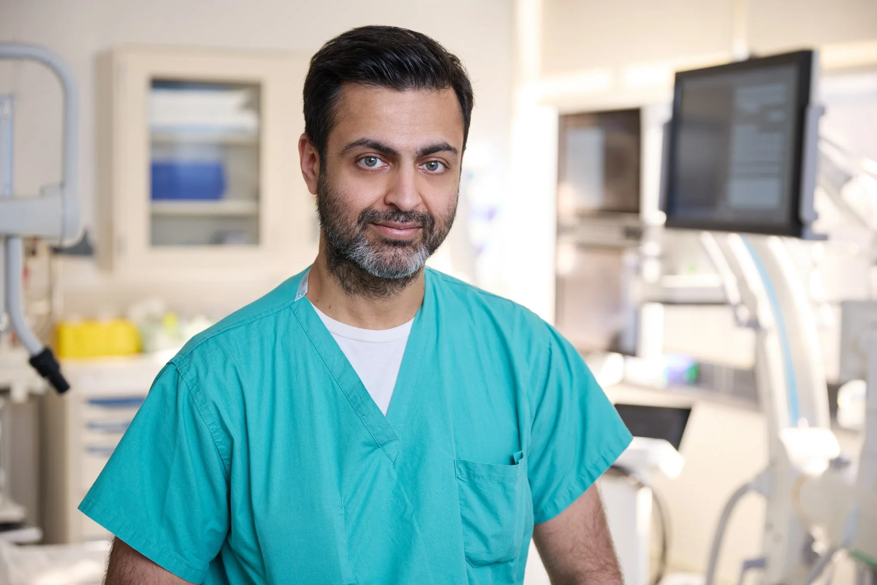 A male healthcare professional of Indian descent Doctor Udit Chaddha with dark hair and a beard, wearing teal medical scrubs, standing in a hospital or clinical setting with medical equipment and monitors in the background. At Mount Sinai Hospital in