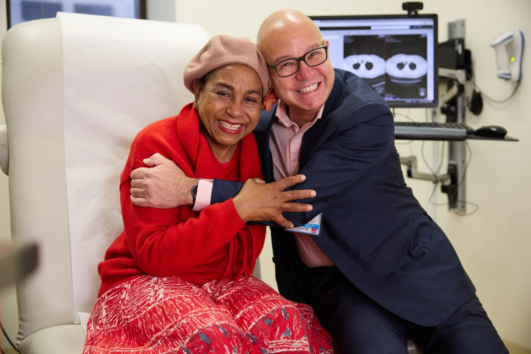 A woman in a red sweater and pink patterned pants sitting in a hospital bed, hugging a smiling surgeon Doctor Andrew Kaufman in a navy blazer and glasses, with medical imaging on a screen behind them. At Mount Sinai Hospital in New York City