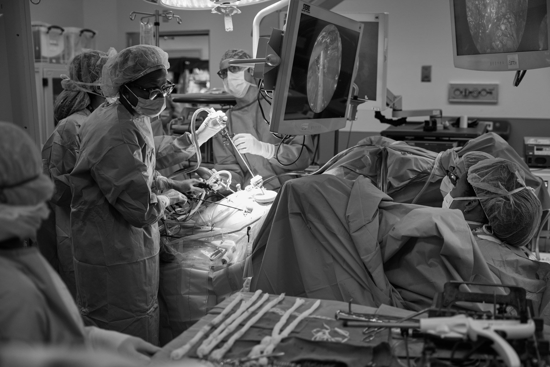 Black and white photo of a medical team led by Dr Patricia Sylla and Dr Sue Hahn performing gastrointestinal surgery in an operating room with monitors displaying the surgical site. At Mount Sinai hospital in New York City