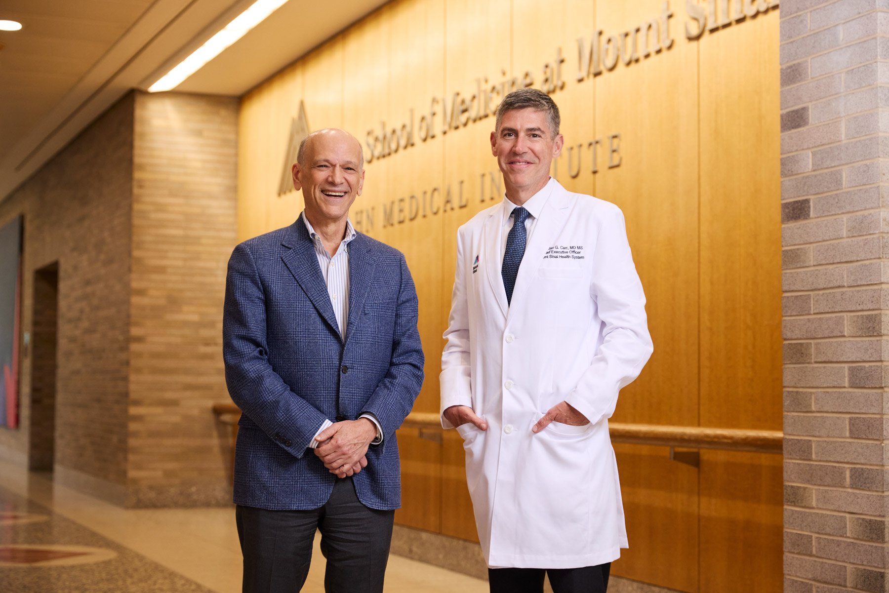 Two men Dr Eric Nestler and Dr Brendon Carr standing in a hospital or medical building lobby at Mount Sinai in New York City. One is wearing a blue blazer and the other in a white medical coat, smiling at the camera.