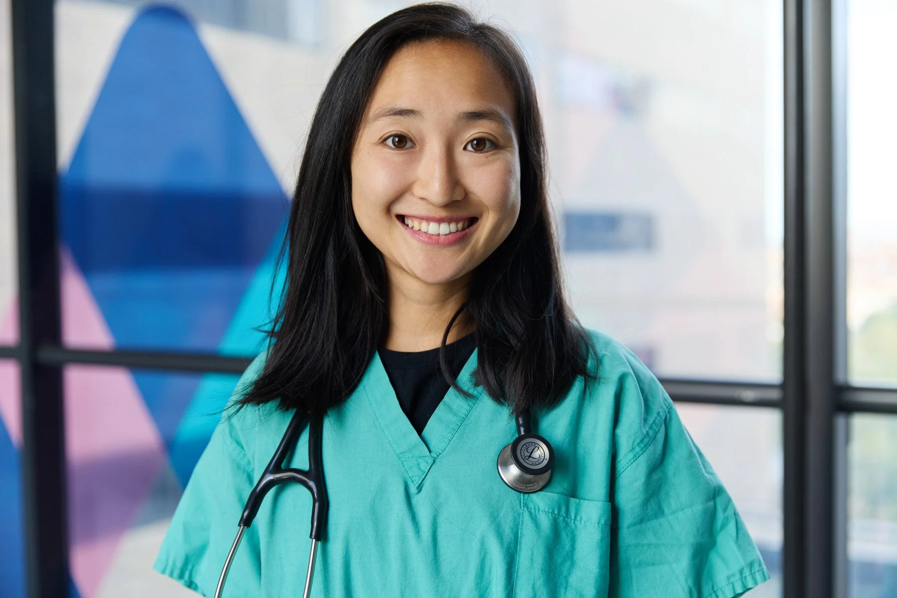 A young female Asian healthcare professional Joy Jiang smiling, wearing teal scrubs with a stethoscope around her neck, standing indoors near large windows with the colorful geometric design of Mount Sinai outside. At Mount Sinai hospital in New York