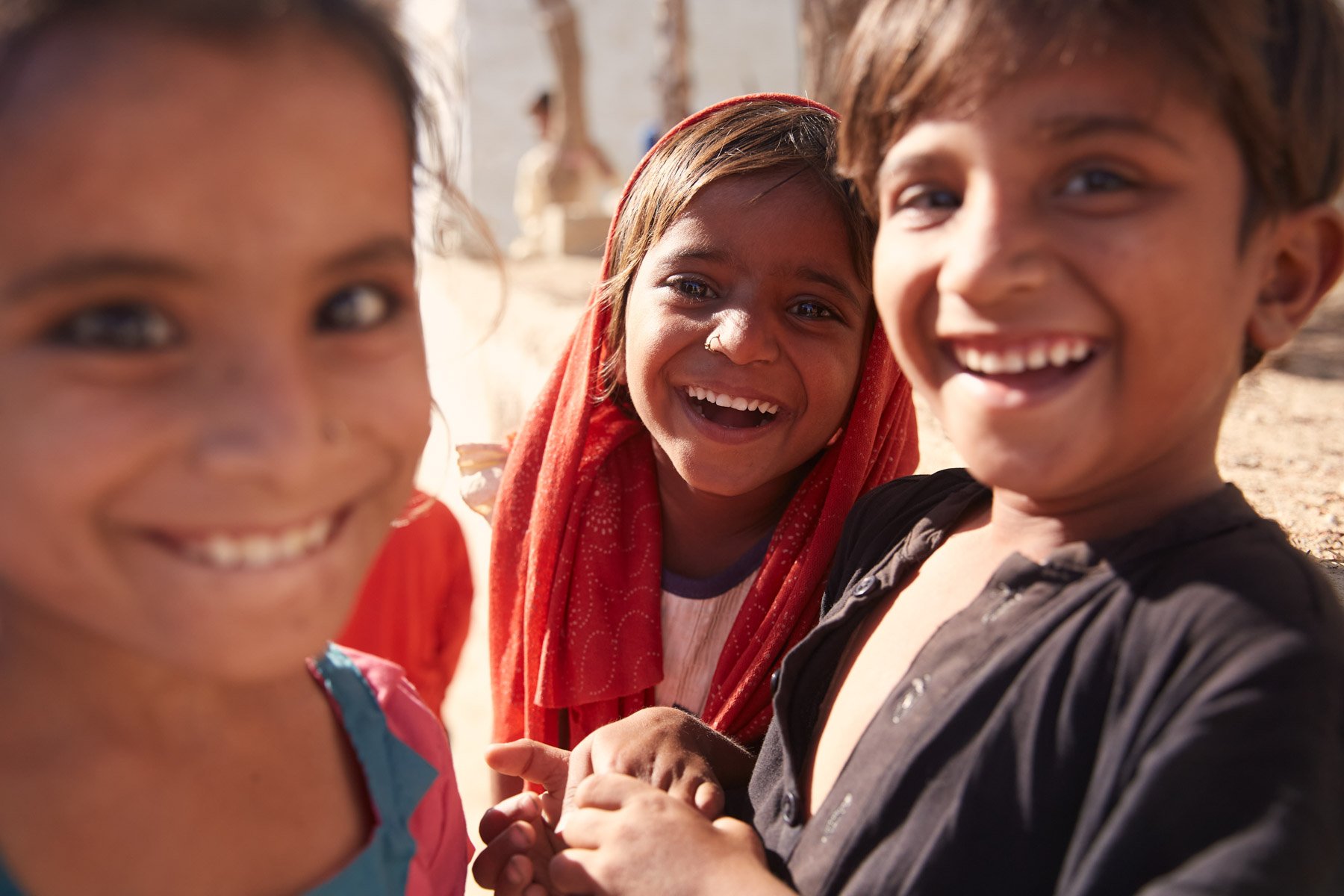 Smiling children playing outdoors in a sunny environment, holding hands and wearing colorful clothing. For Teach the World Foundation, Razzaqabad, Karachi, Pakistan.