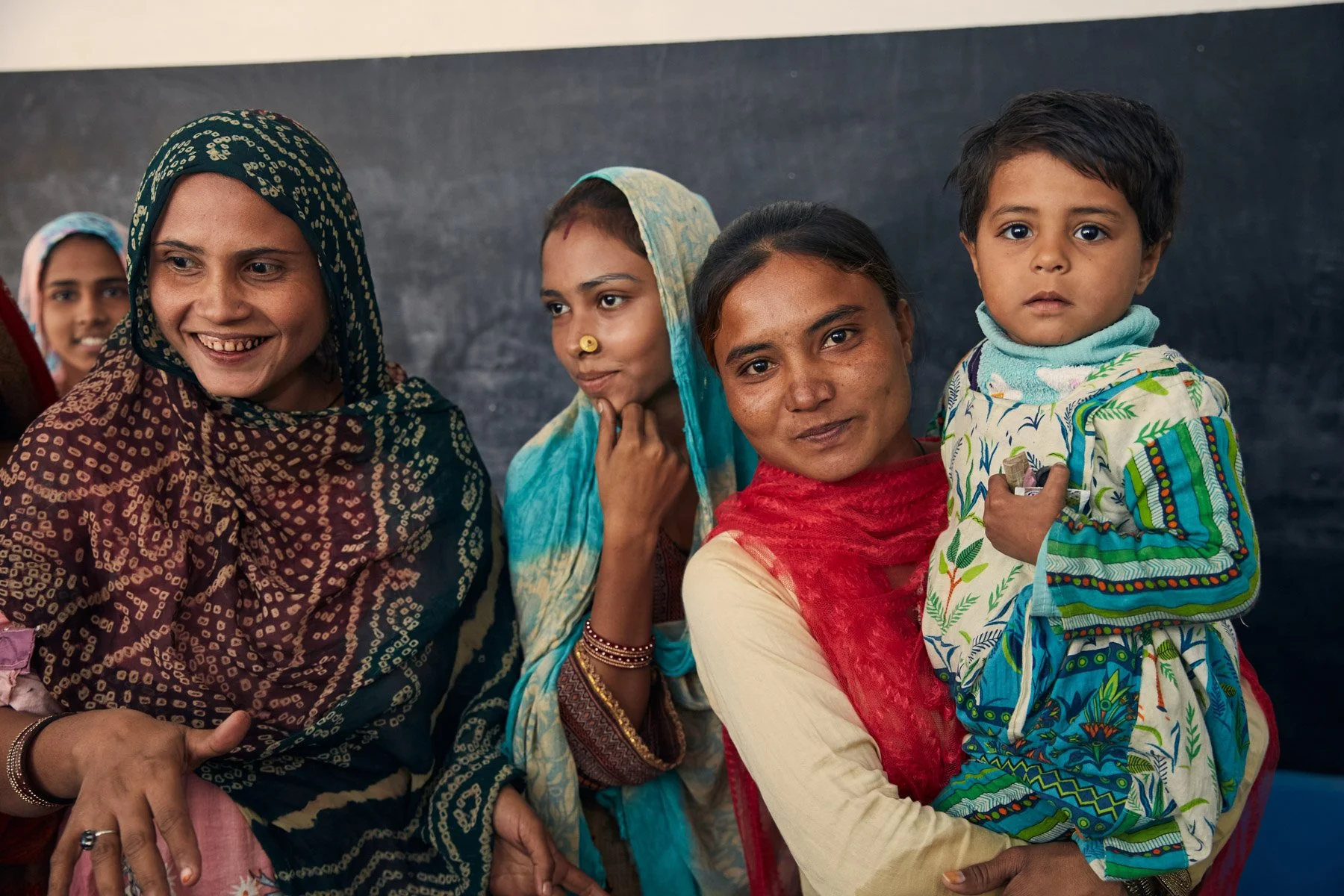 Group of five Pakistani women and children beside a blackboard, wearing colorful traditional clothing, smiling and looking at the camera. For Teach the World Foundation, Razzaqabad, Karachi, Pakistan.