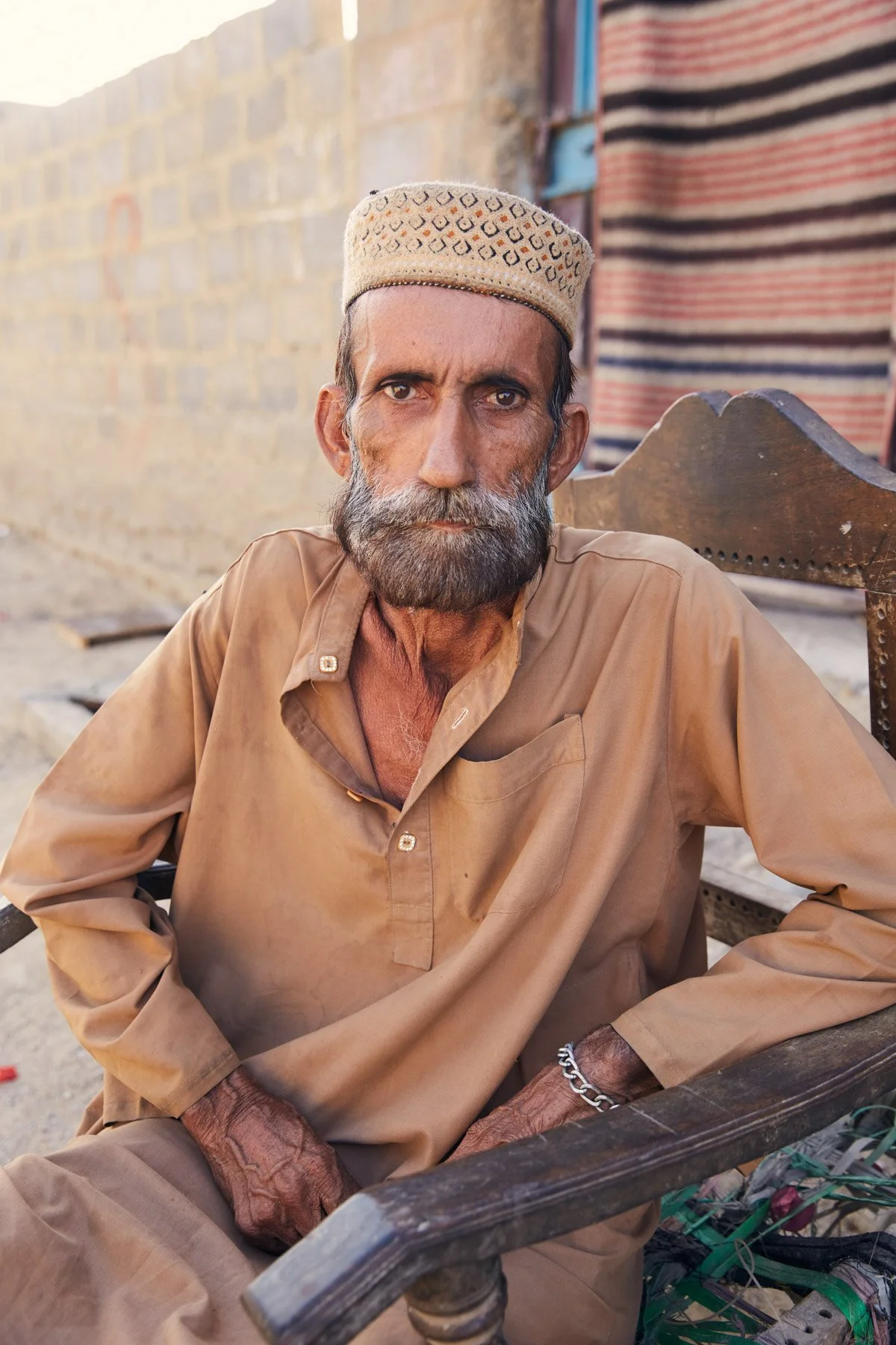 An elderly man with a beard and mustache, wearing a traditional cap and tan shirt, sitting on a wooden chair outdoors in front of a brick wall, looking directly at the camera. For Teach the World Foundation, Razzaqabad, Karachi, Pakistan.