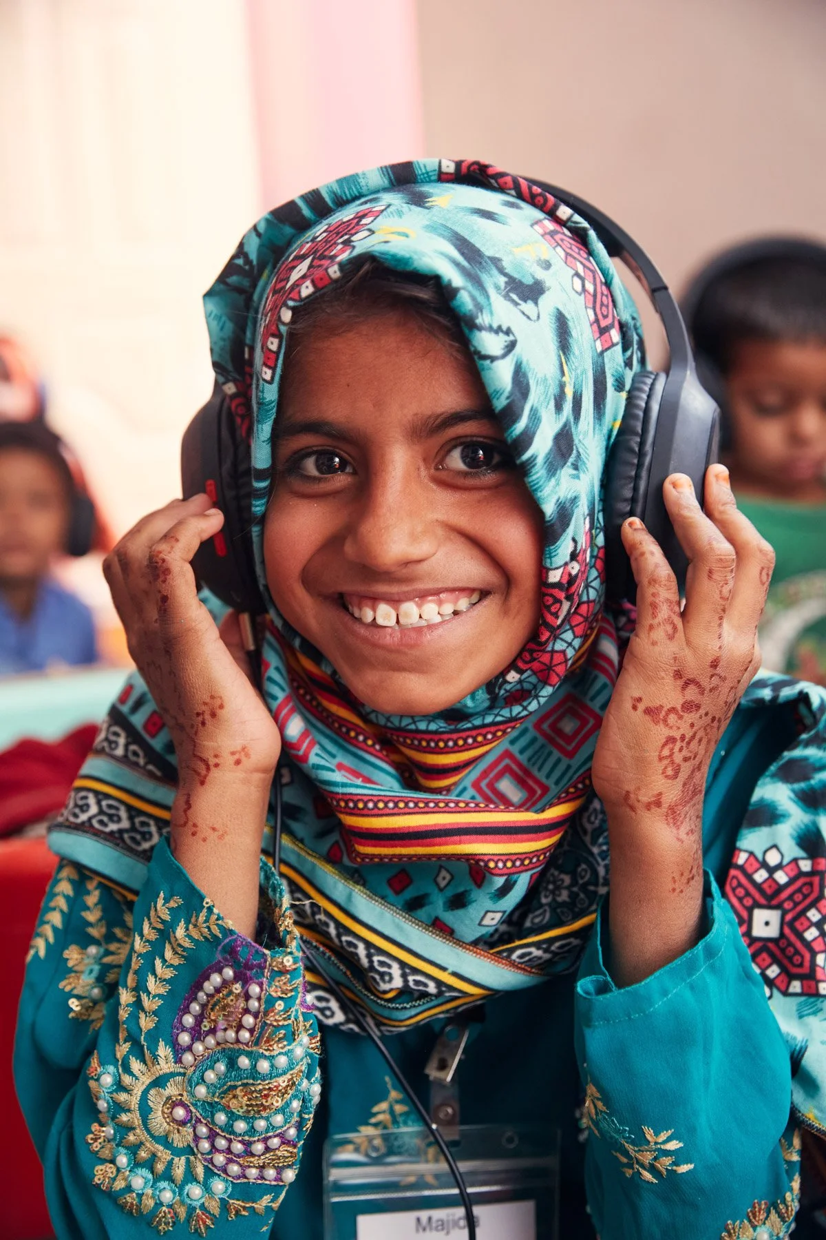 A young girl smiling wearing headphones and a colorful hijab, with henna on her hands. For Teach the World Foundation, Razzaqabad, Karachi, Pakistan.