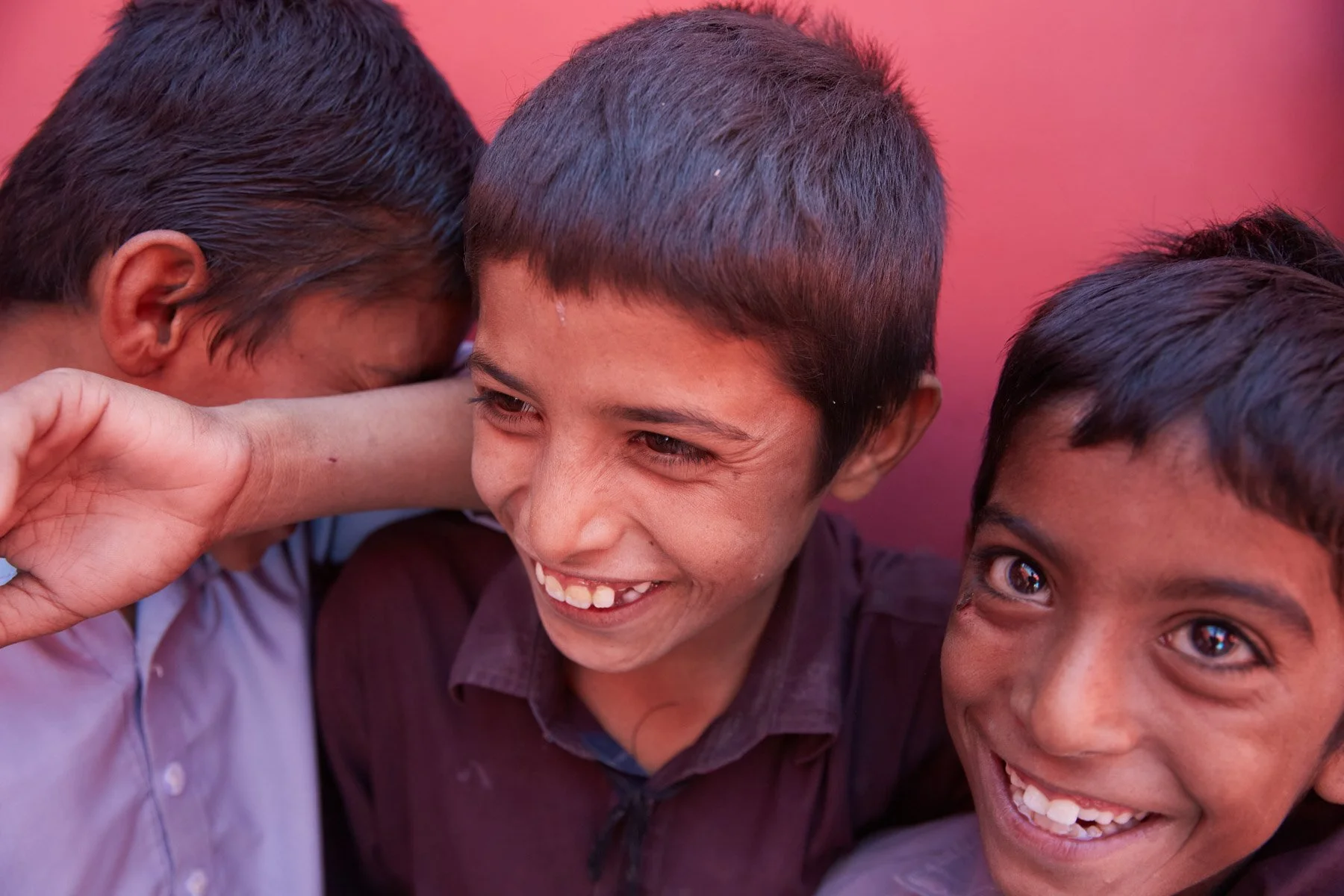 Three young boys smiling and laughing closely together, with one boy covering his ear. For Teach the World Foundation, Razzaqabad, Karachi, Pakistan.