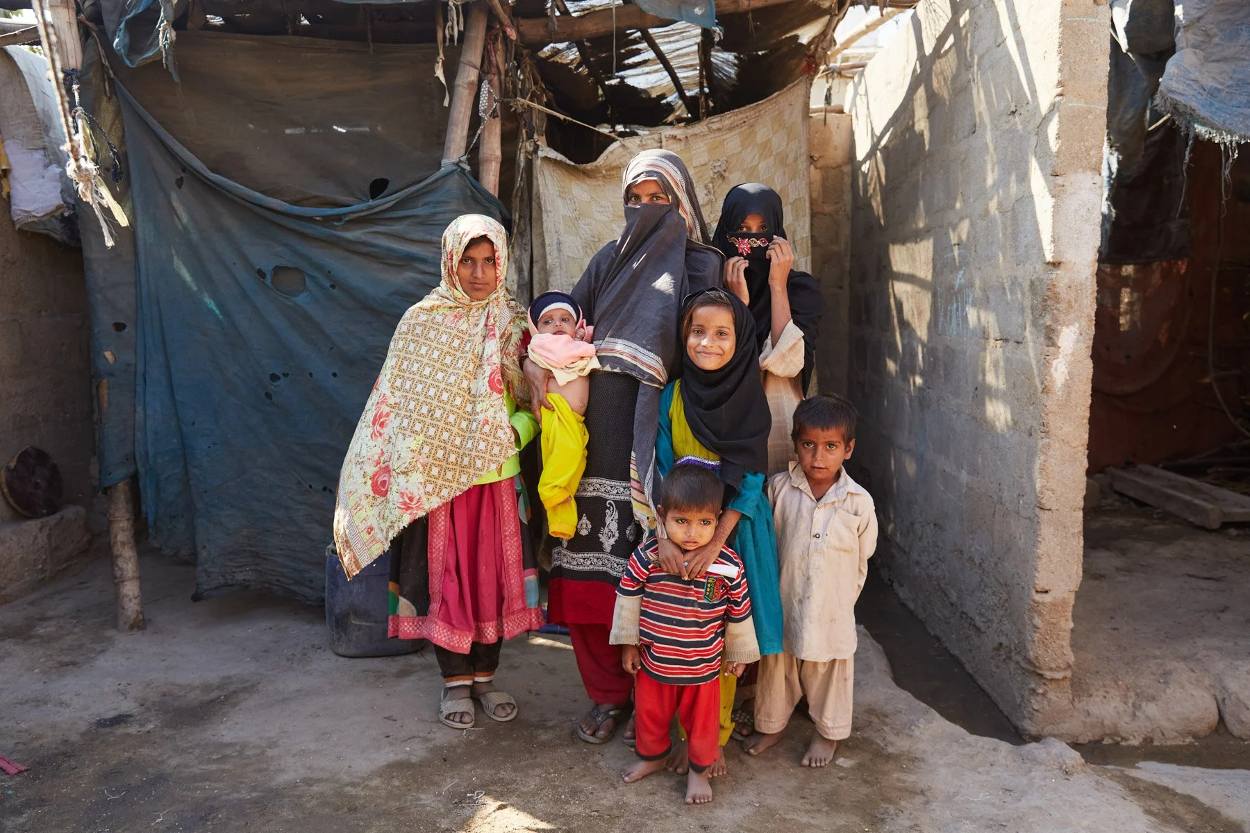 A family standing outside of a makeshift shelter made of fabric and wood, in a rural or impoverished area. For Teach the World Foundation, Razzaqabad, Karachi, Pakistan.